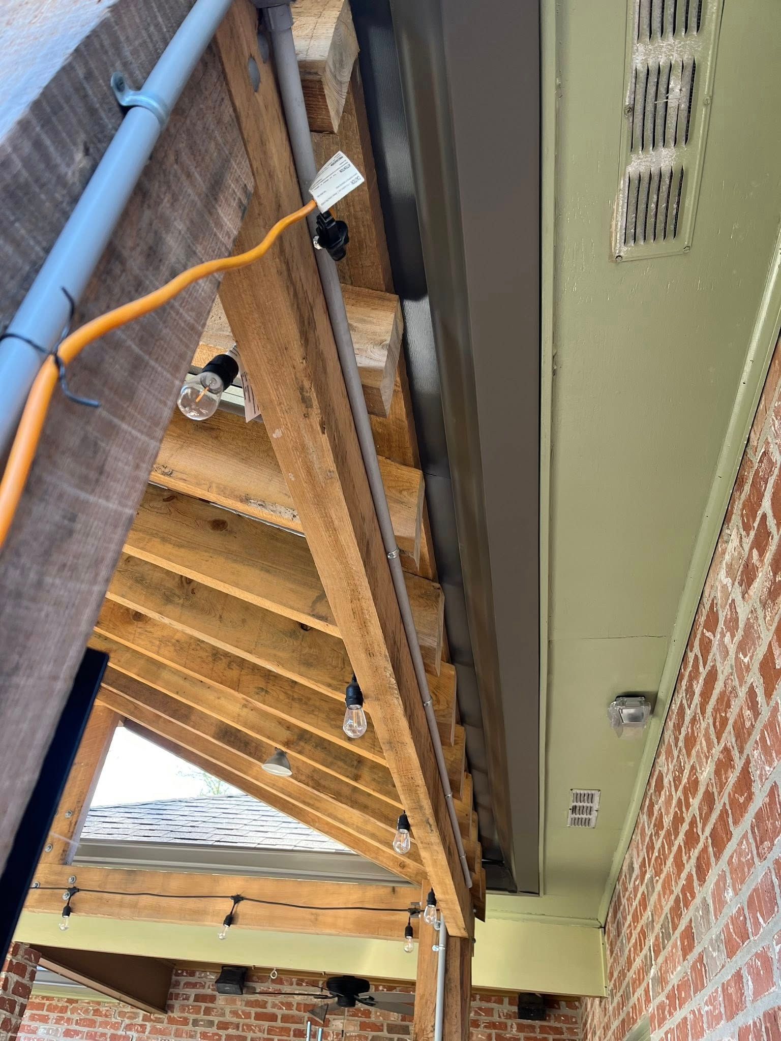 Upward view of porch roof framing with exposed wooden beams and green trim beside brick wall
