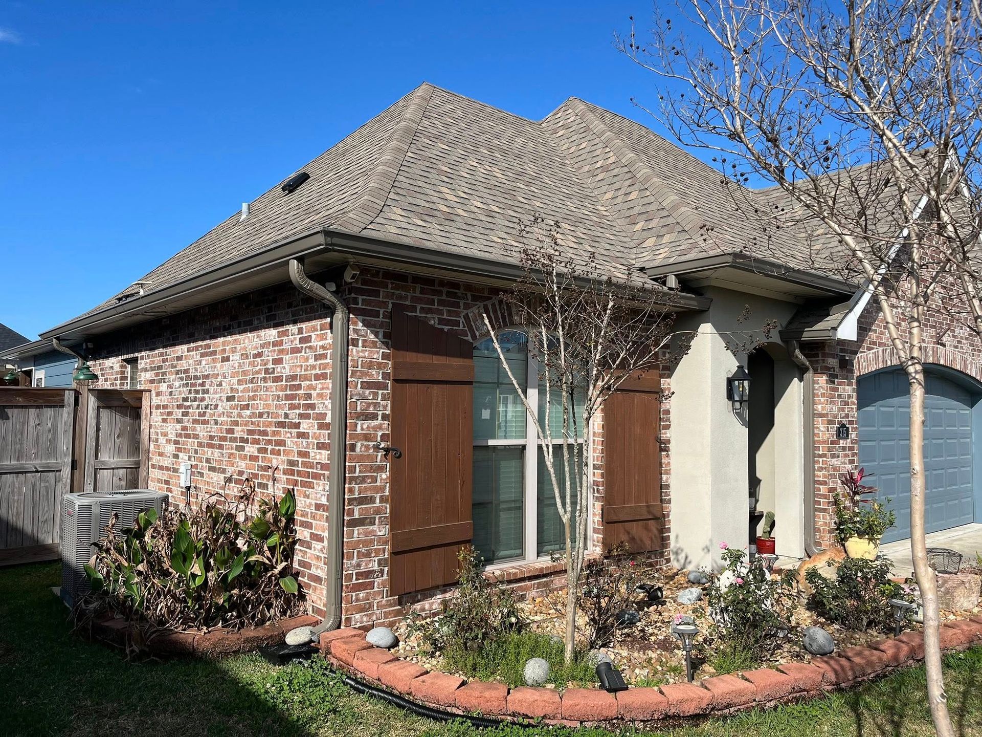 Brick house exterior with gray shingled roof, wood shutters, and a small landscaped front garden under a blue sky