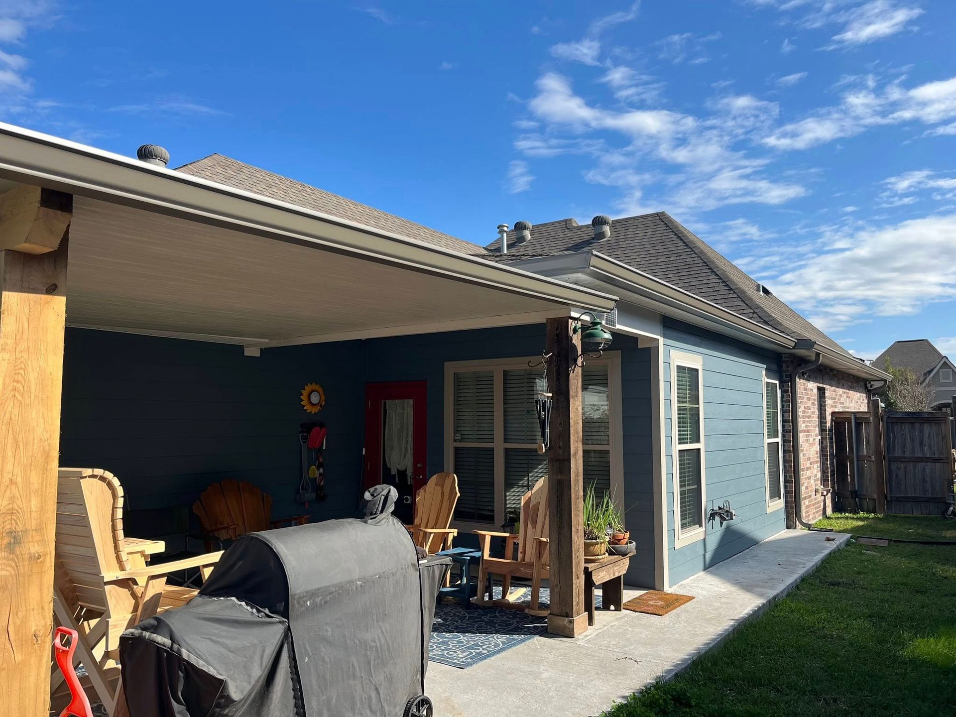 Covered backyard patio with grill, seating, and blue house exterior under a sunny sky.