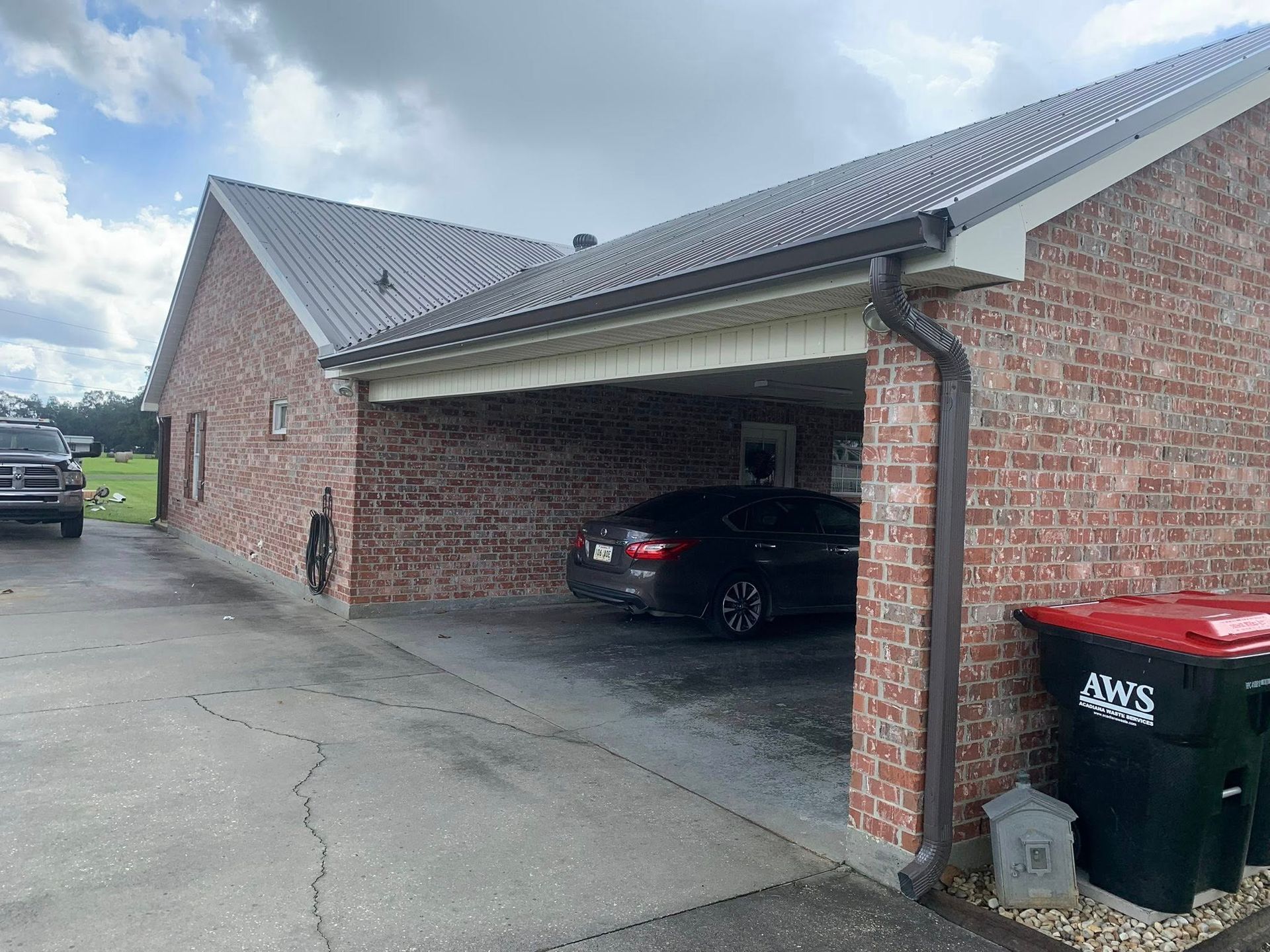 Brick house carport with a black car, trash bins, and a driveway under a cloudy sky