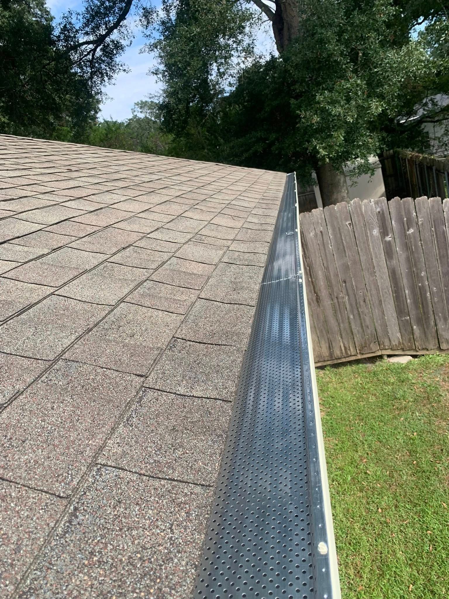 House roof edge with gutter and leaf guard beside a wooden fence and green yard