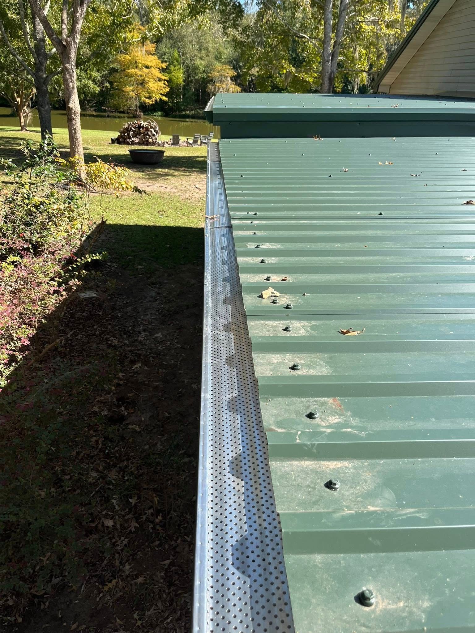 Green metal roof with gutter and perforated edge beside a grassy yard and trees