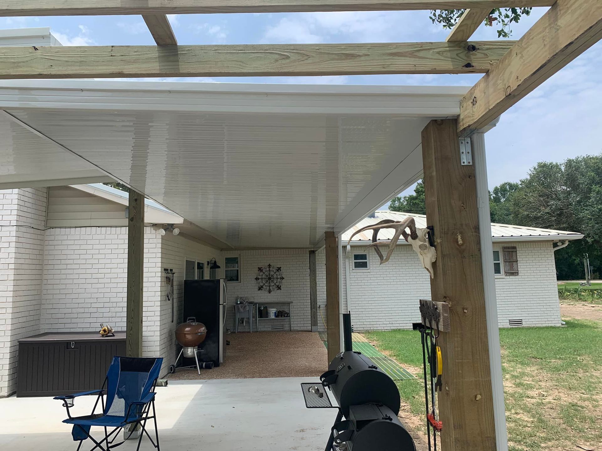 Covered patio with grill, chairs, and brick flooring beside a white house and grassy yard