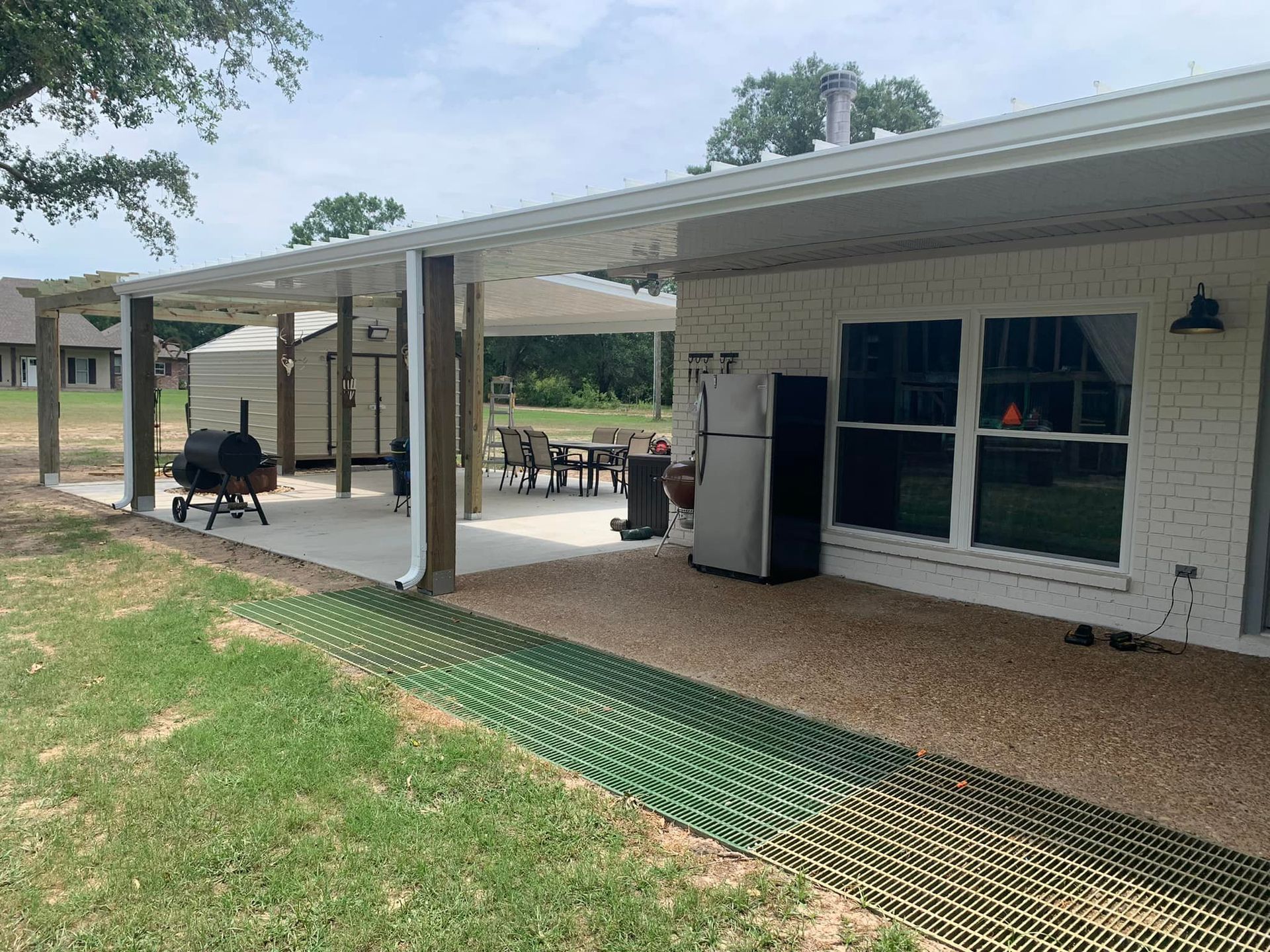 Covered patio with outdoor seating beside a white brick house and grassy yard