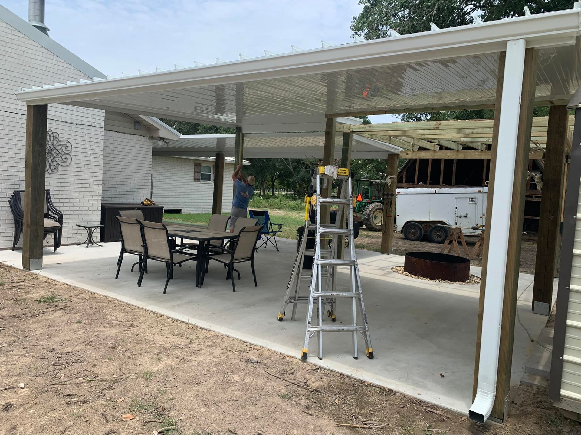 Covered patio with white roof, patio table and chairs, and a ladder on a concrete slab beside a house