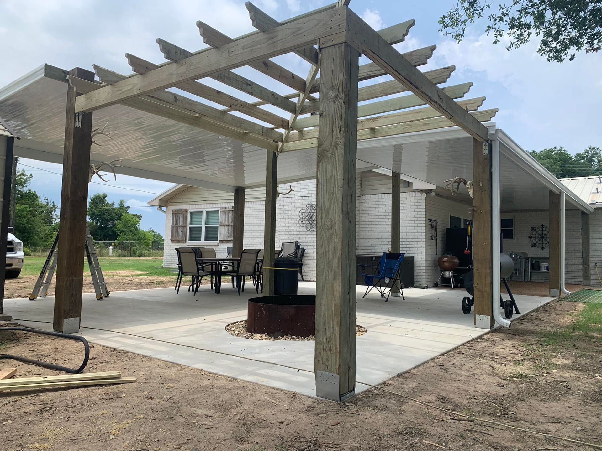 Backyard patio with pergola roof, concrete floor, chairs, grill, and a house in the background