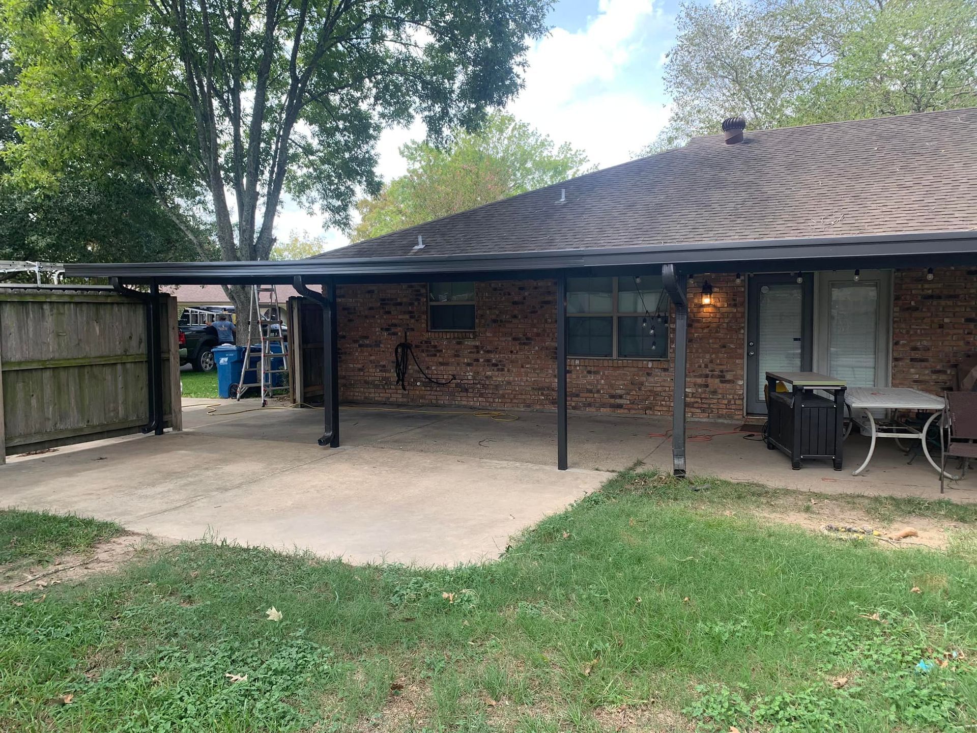 Backyard patio beside a brick house with a covered carport, concrete slab, and outdoor furniture.