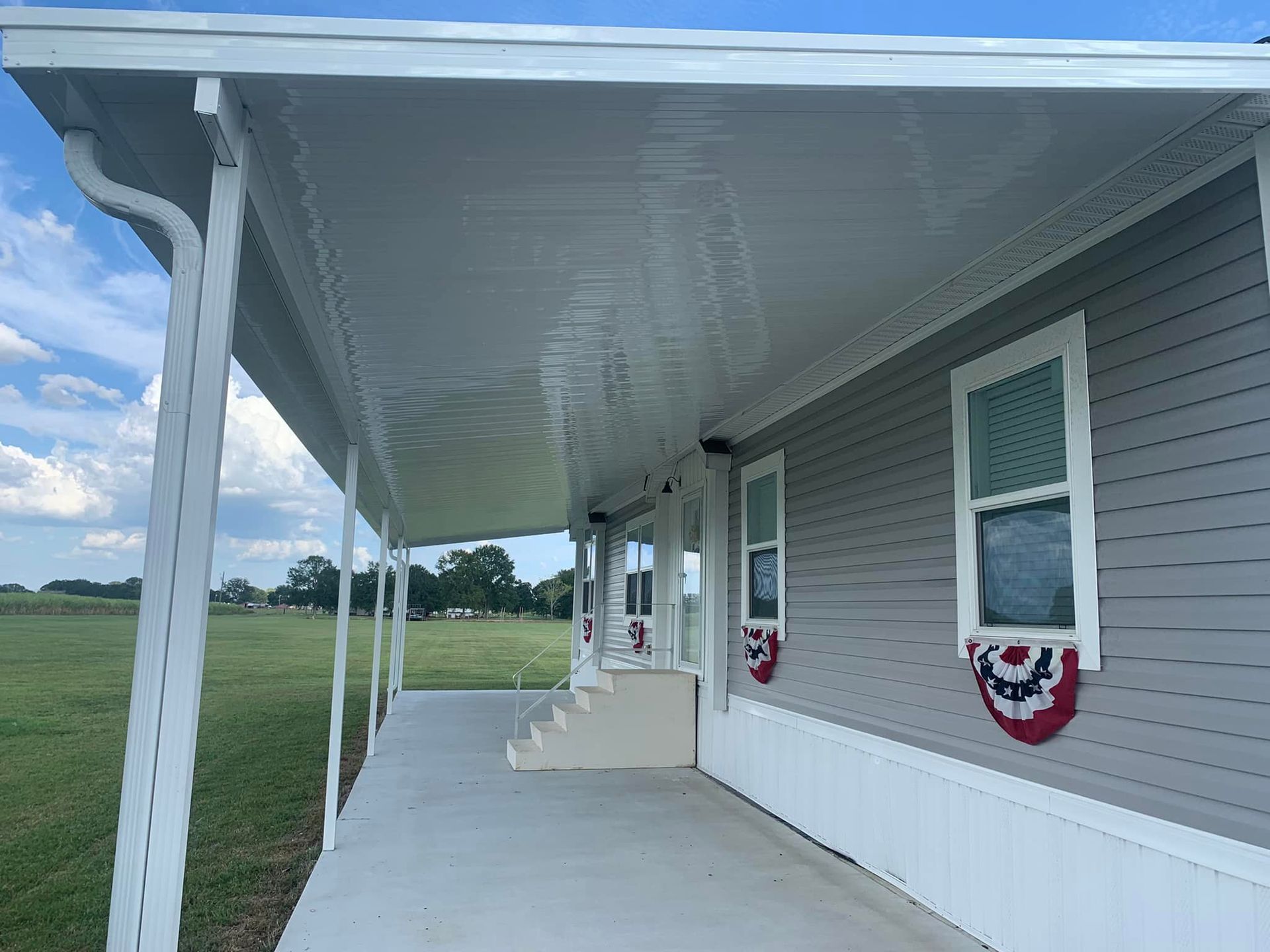 Covered porch with white siding and columns beside a grassy field under a blue sky.