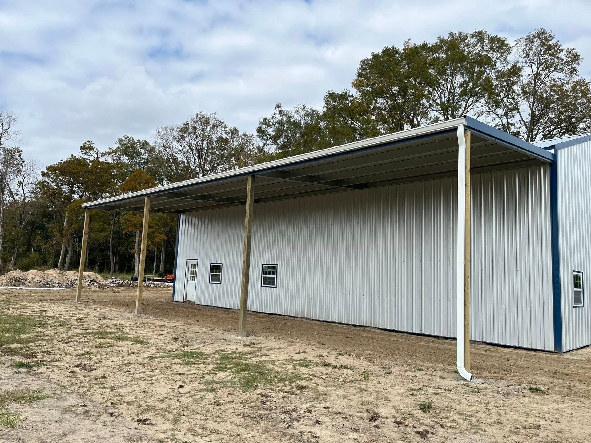 White metal shed with a covered porch on a bare dirt lot, surrounded by trees under a cloudy sky