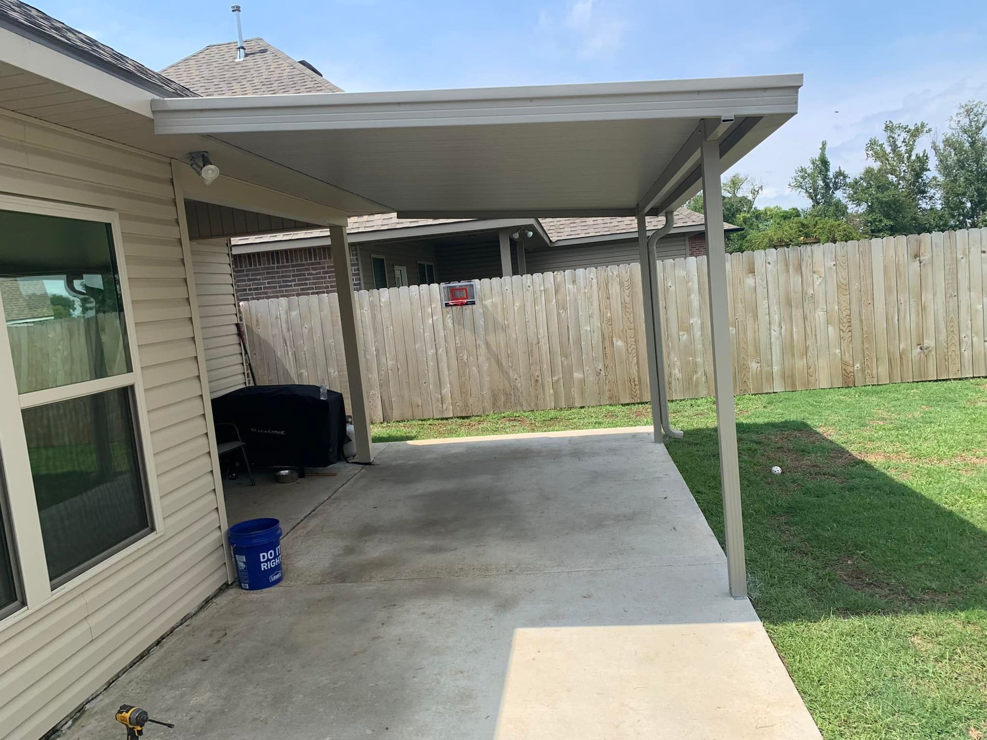 Covered concrete patio beside a house, with a white fence and green lawn in the backyard.