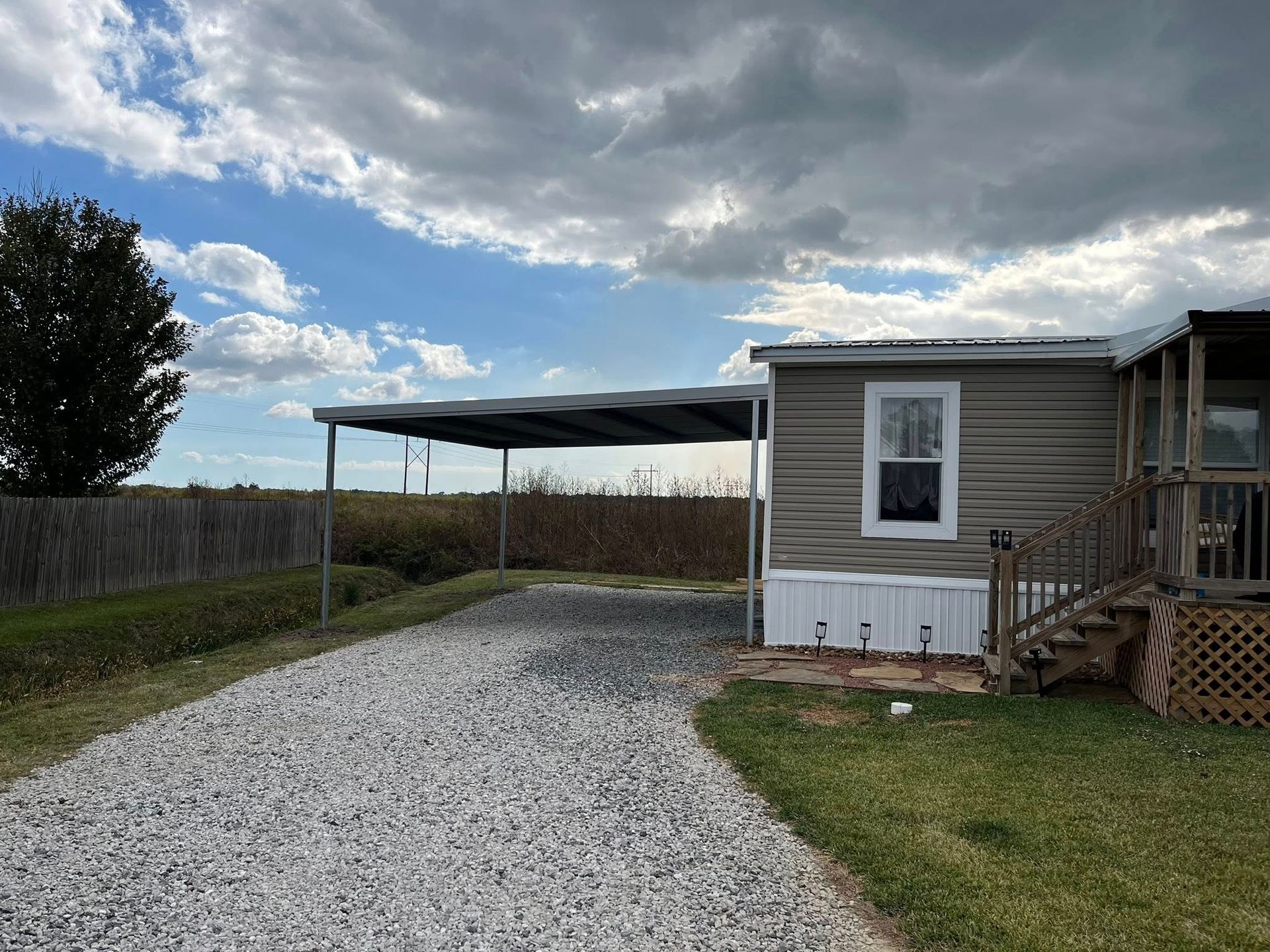 Gravel driveway leading to a beige house with a carport under a cloudy sky