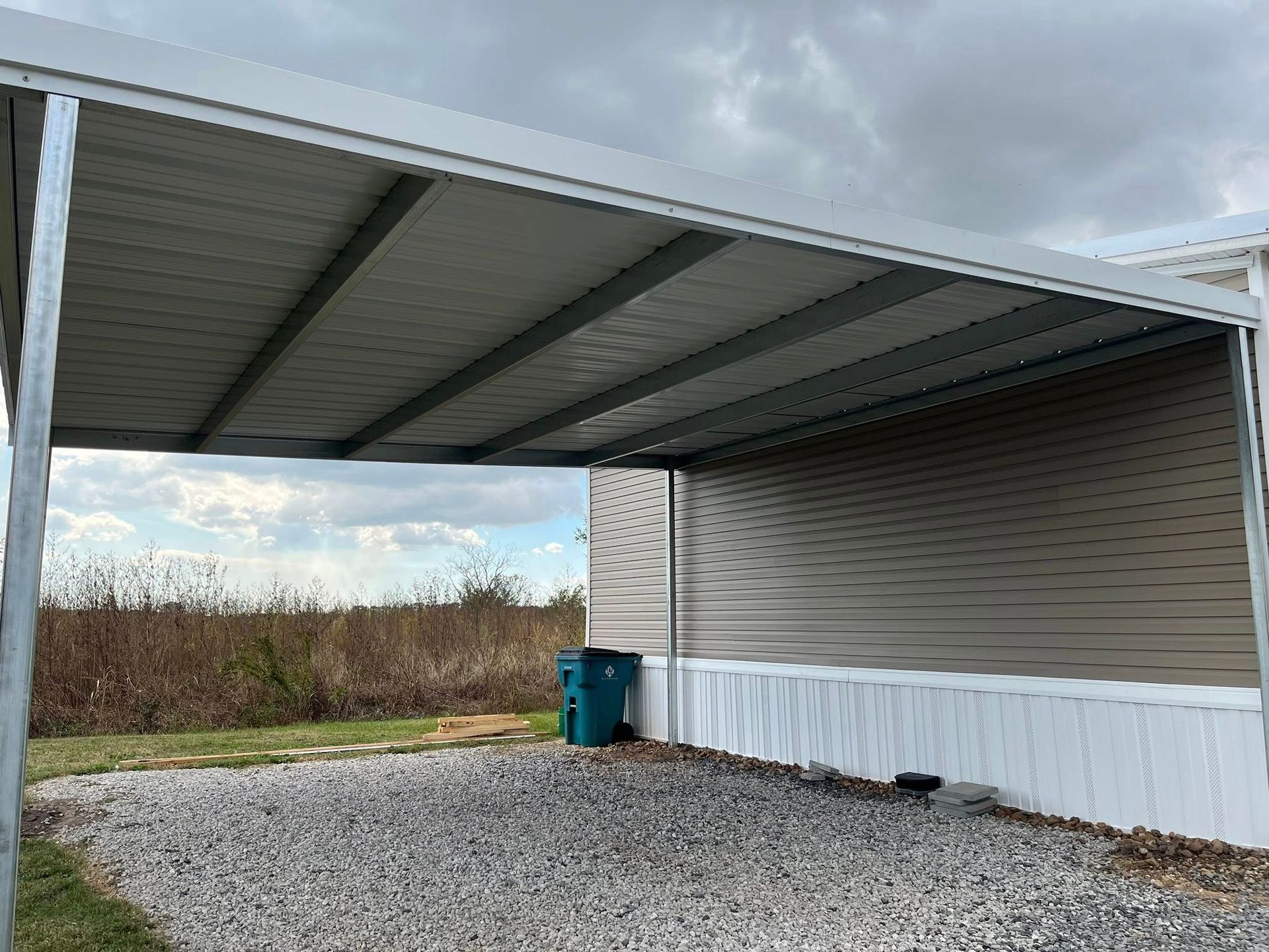Metal carport attached to a gray building over gravel, with a blue trash bin nearby and cloudy sky beyond