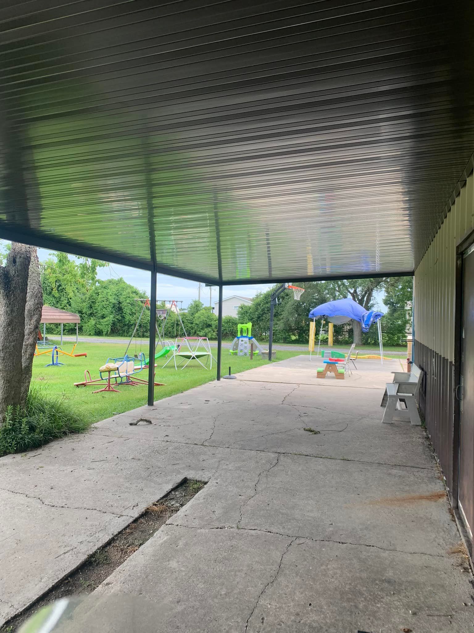Covered concrete walkway beside a grassy park, with a blue canopy and picnic tables in the distance