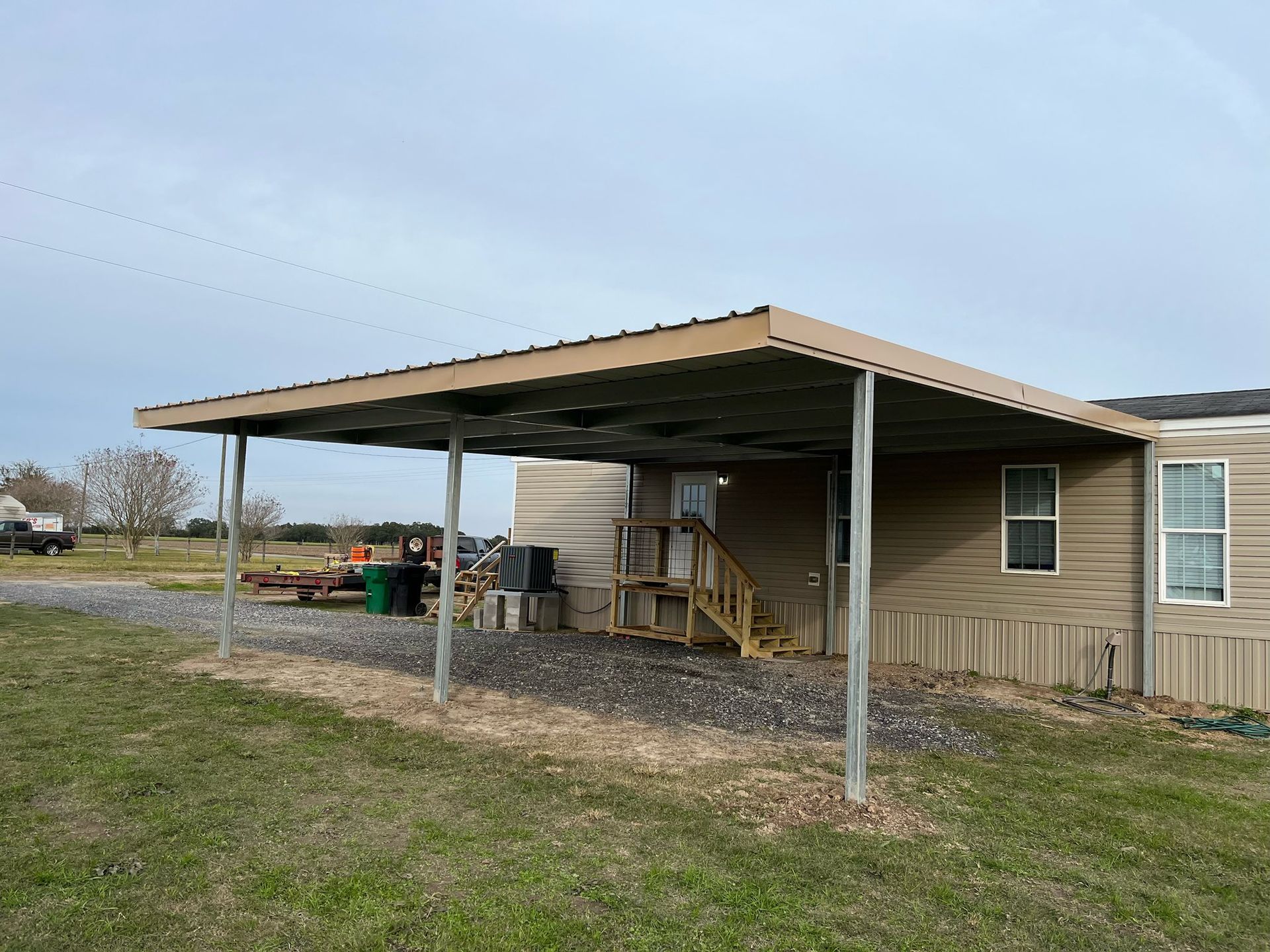 Large tan house with a covered carport, stairs, and outdoor items in a grassy yard under cloudy sky