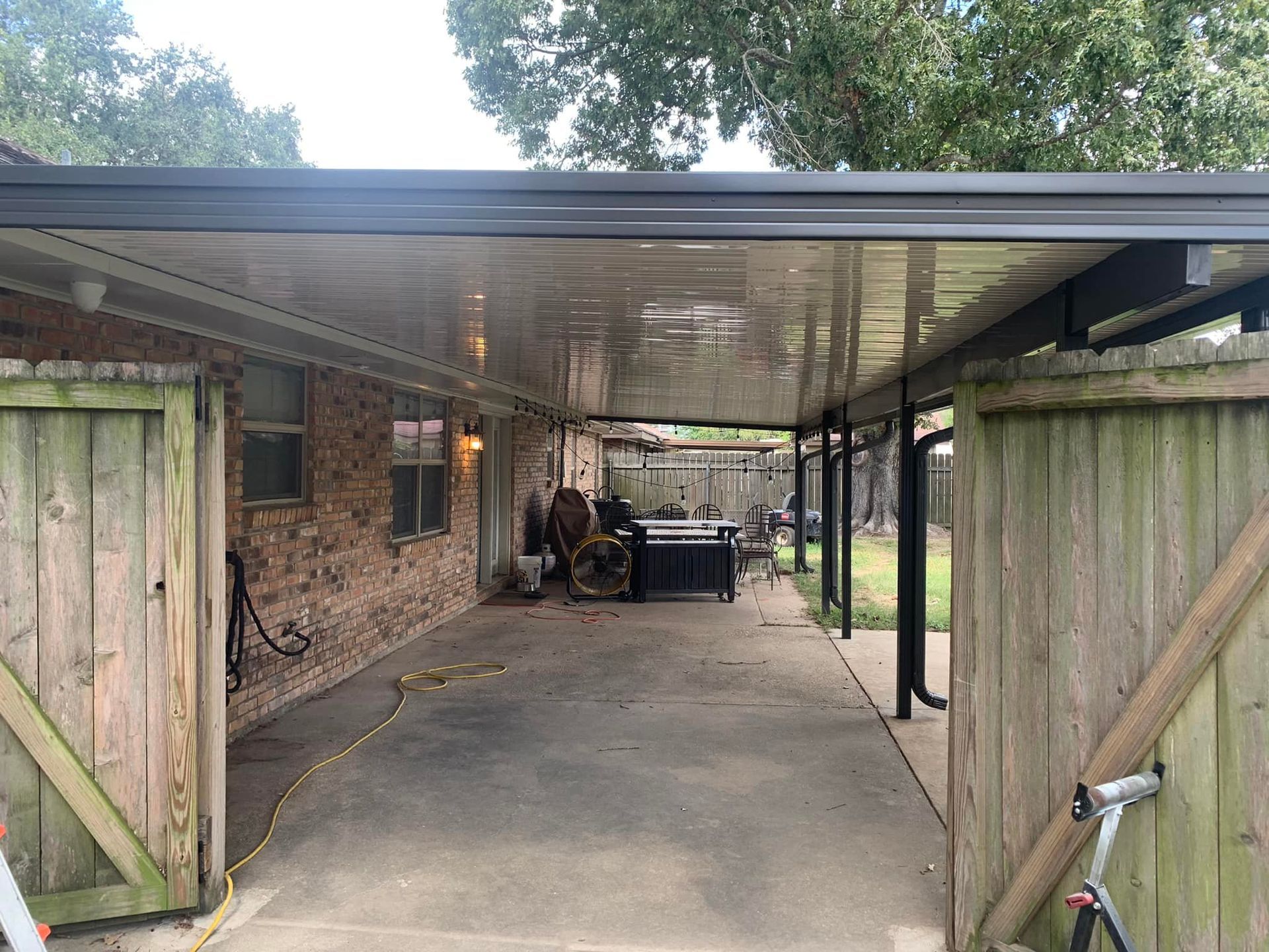 Covered driveway/carport beside a brick house, with a concrete path and a few tools at the far end