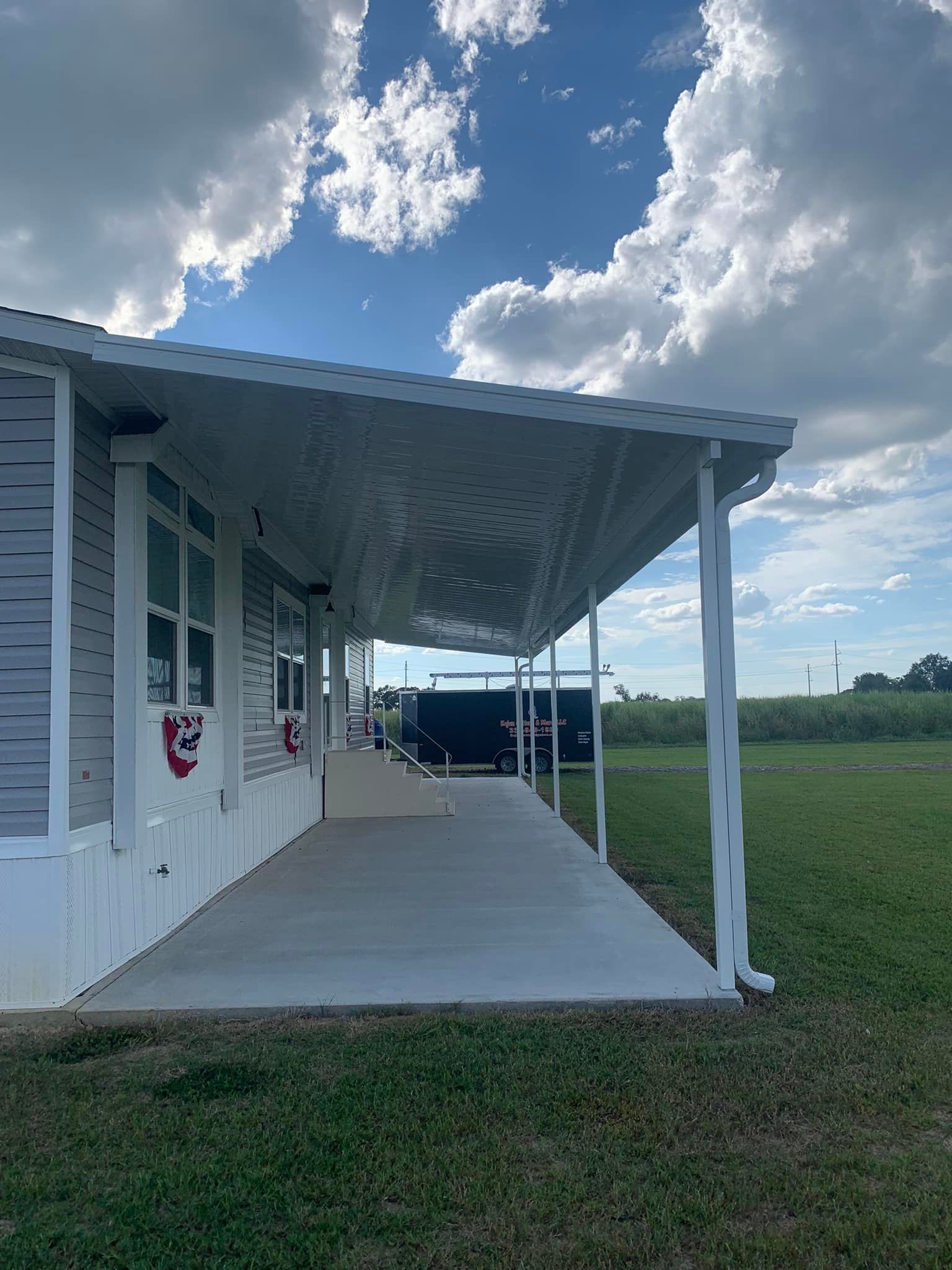 Covered porch of a white building with a concrete ramp, grassy field, and cloudy blue sky.