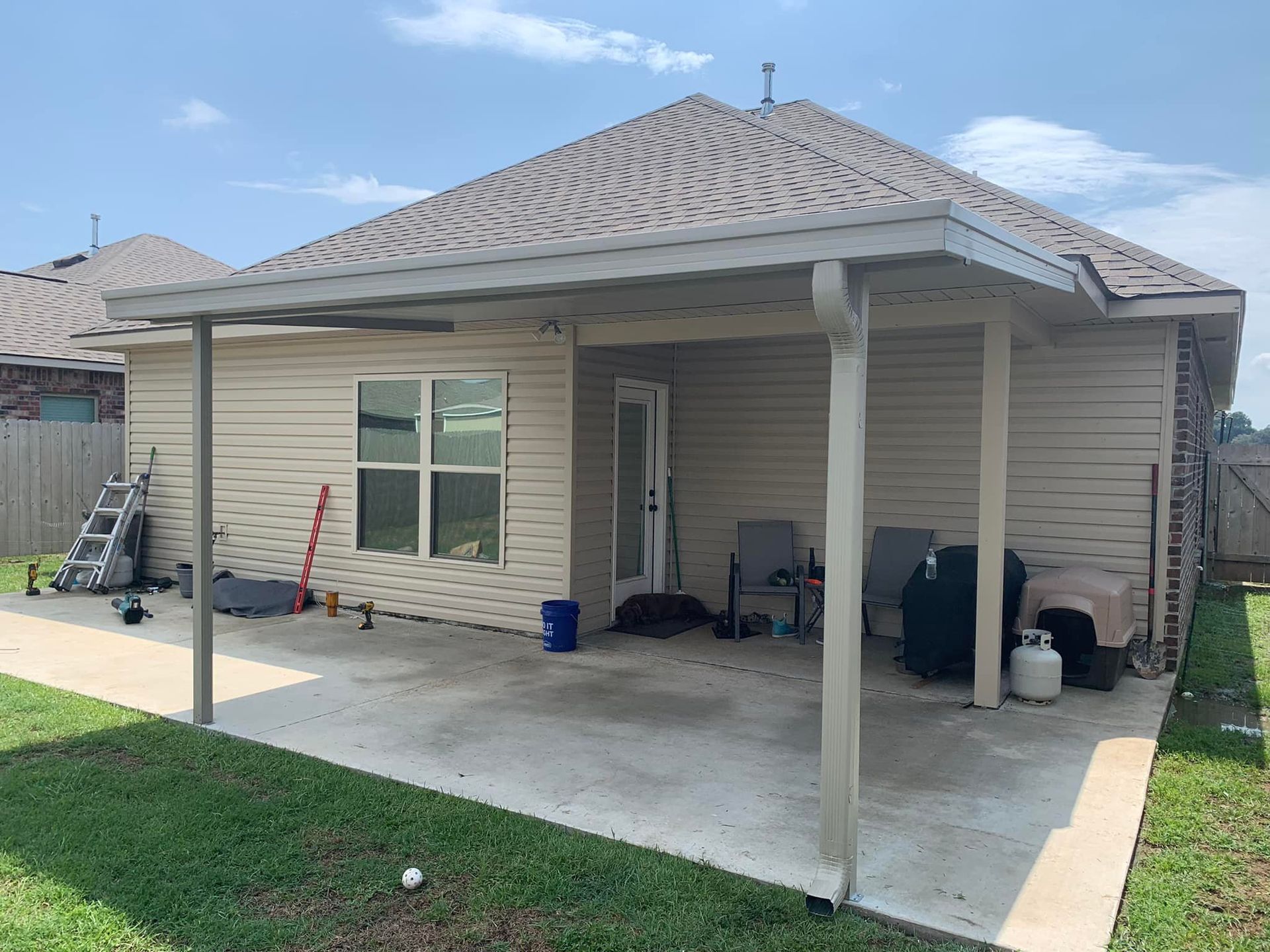 Backyard covered patio with beige siding, white posts, concrete slab, and lawn beyond