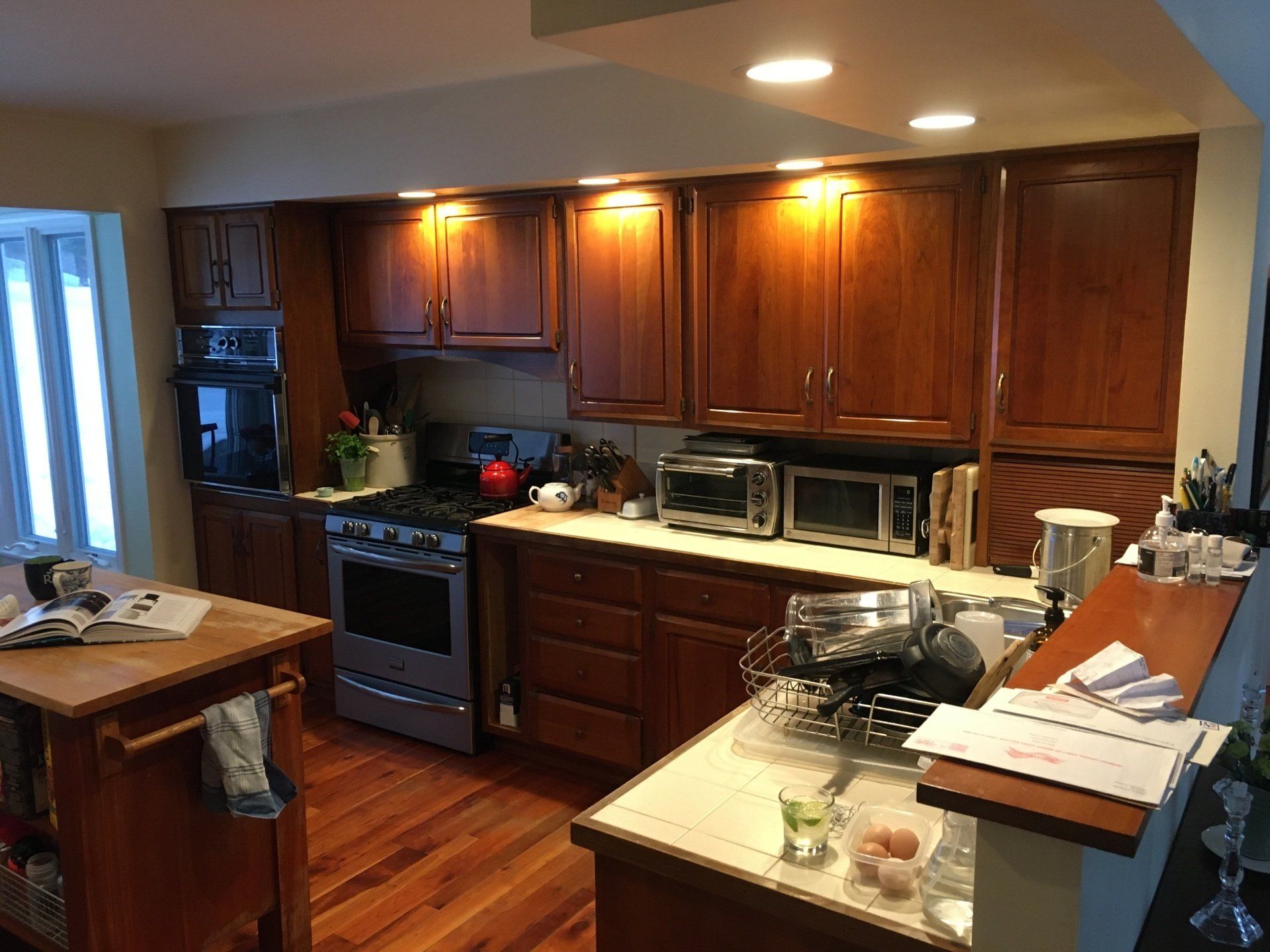 Warm kitchen with wooden cabinets, stainless stove, and a cluttered countertop under recessed lights.