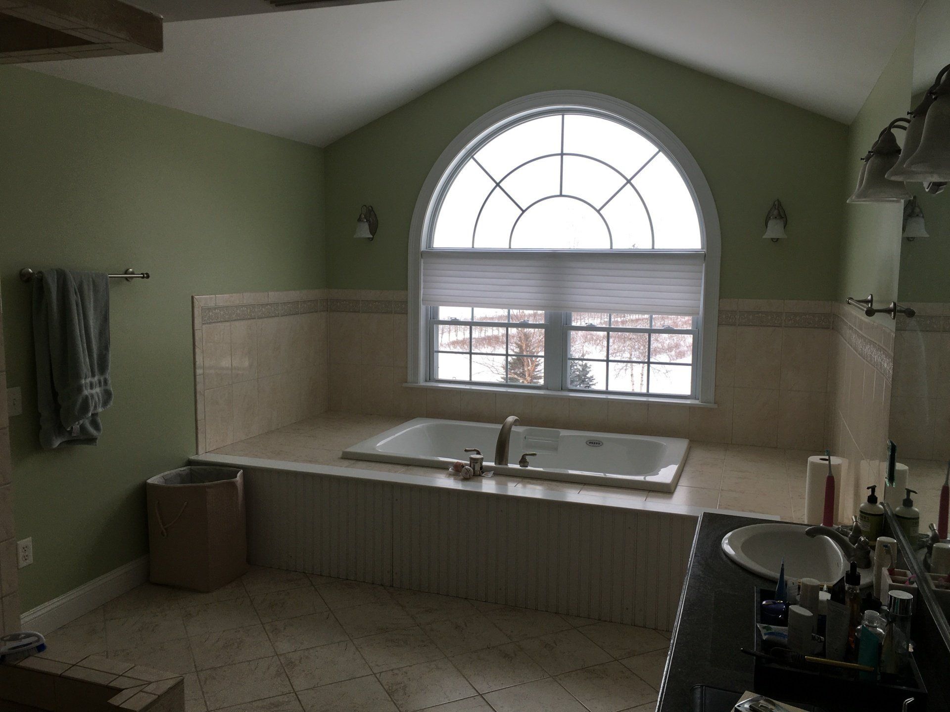 Green bathroom with a large arched window above a double sink and countertop cluttered with toiletries.