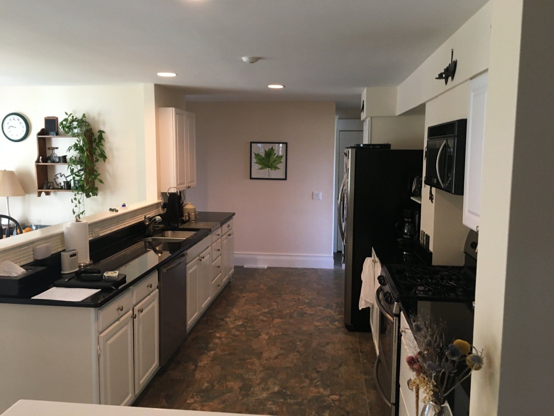 Long galley kitchen with white cabinets, black countertops, and a dark floor, viewed toward a hallway.