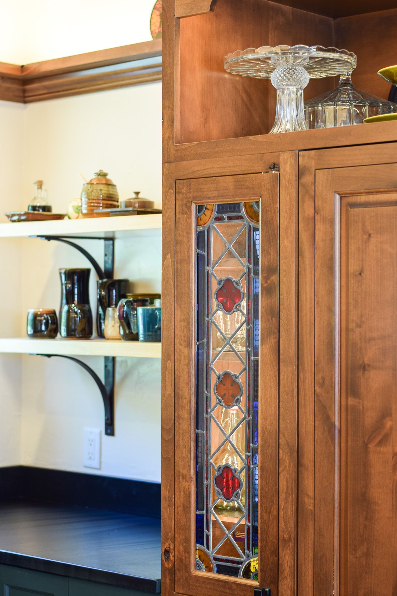 Wooden cabinet with stained-glass detail beside a kitchen shelf holding glassware and small jars