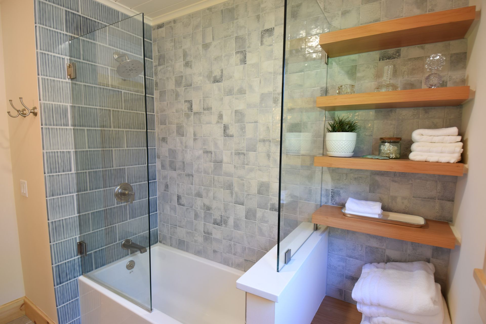 Modern bathroom with glass-enclosed tub, gray tile walls, and wooden shelves holding towels and decor