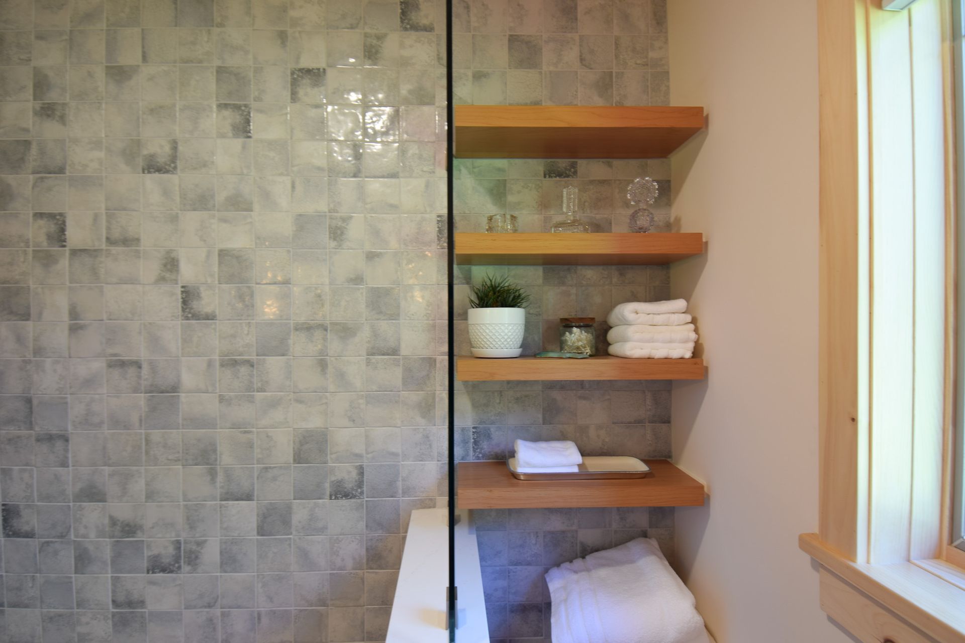 Bathroom with tiled shower wall and wooden shelves holding towels and a small plant