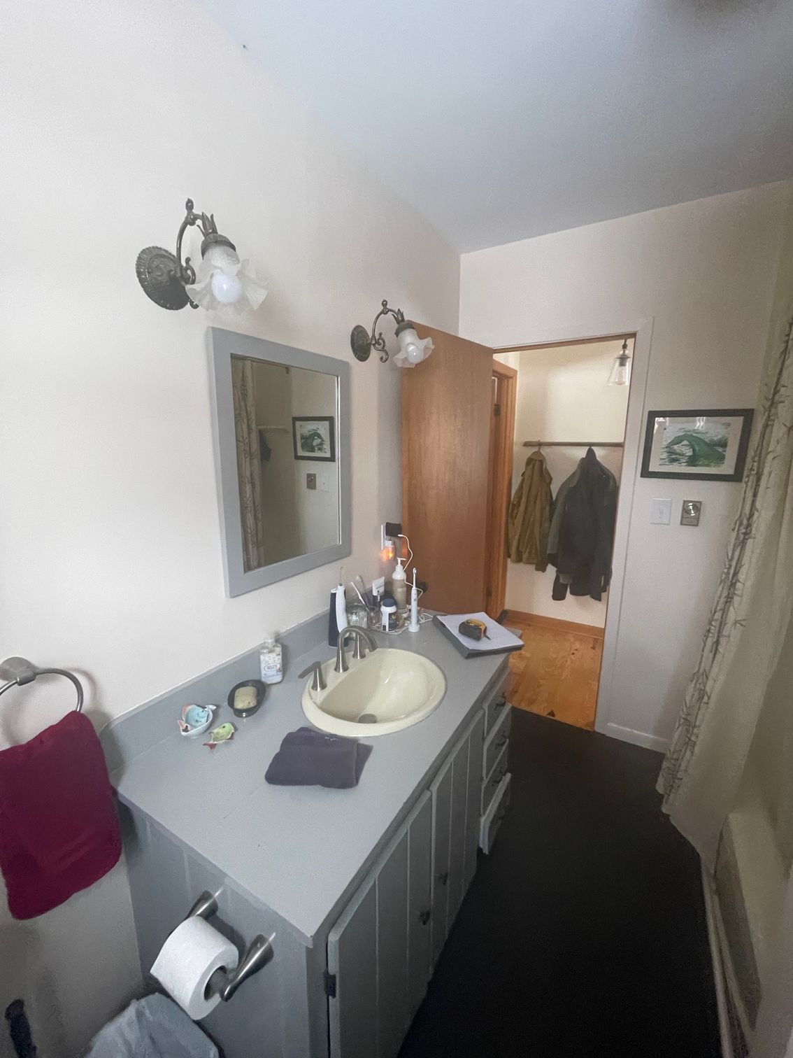 Narrow bathroom with gray vanity, sink, mirror, wall lights, and open door to a hallway with coats.
