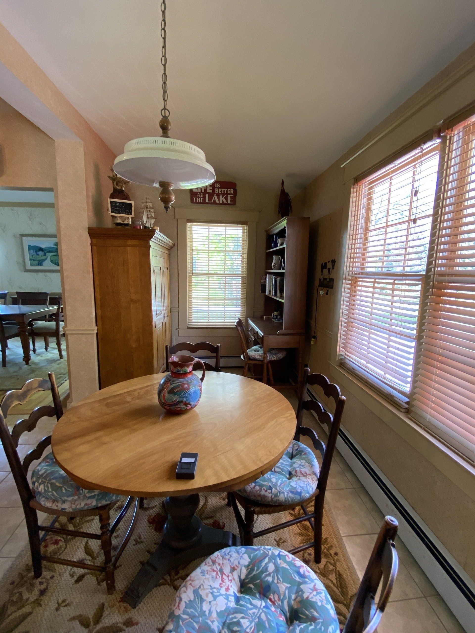 Small dining room with round wooden table, floral chairs, hanging light, and sunlit windows with blinds.
