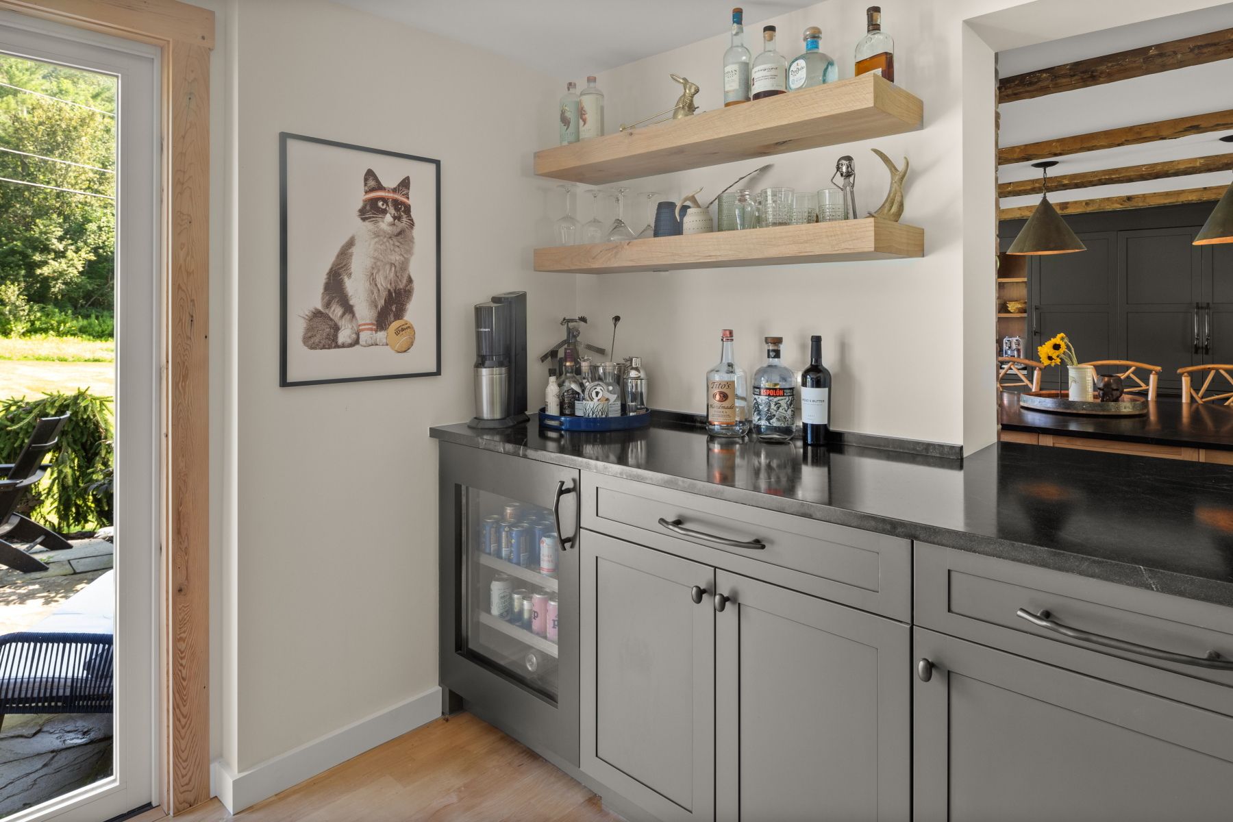 Home bar with gray cabinets, shelves of bottles, a mini fridge, and a framed cat print by an open doorway