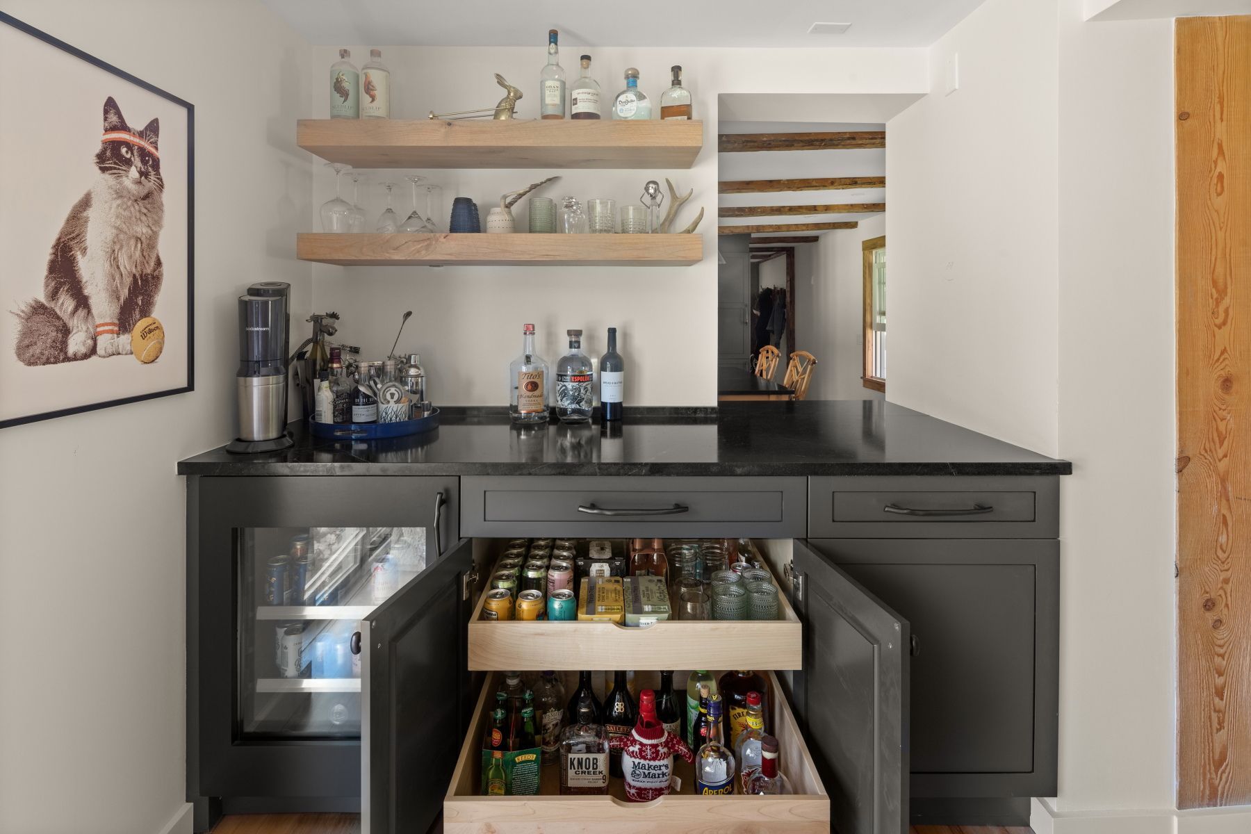 Modern kitchen nook with a black counter, open drawer of snacks, small fridge, and cat artwork on the wall