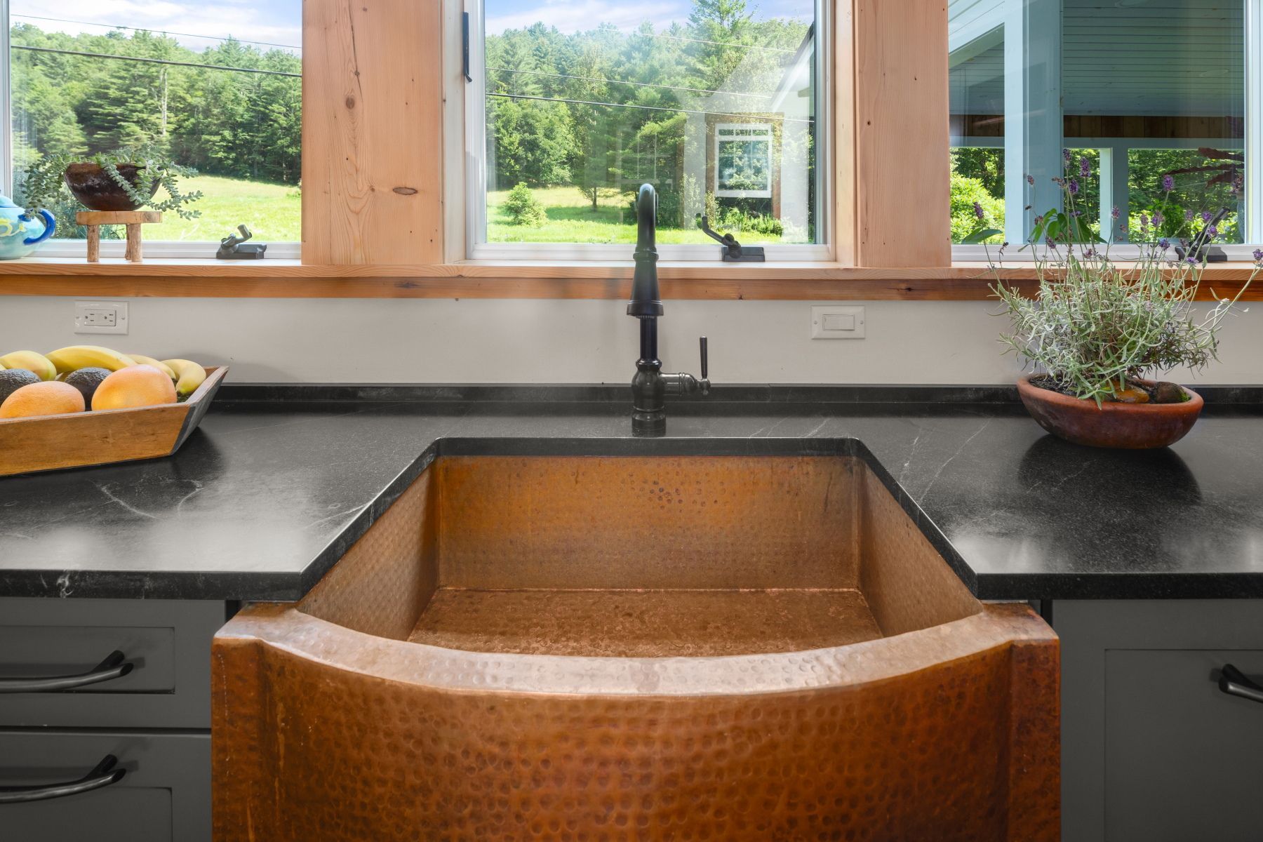 Modern kitchen sink with copper basin under windows, black countertops, and fruit bowls on either side