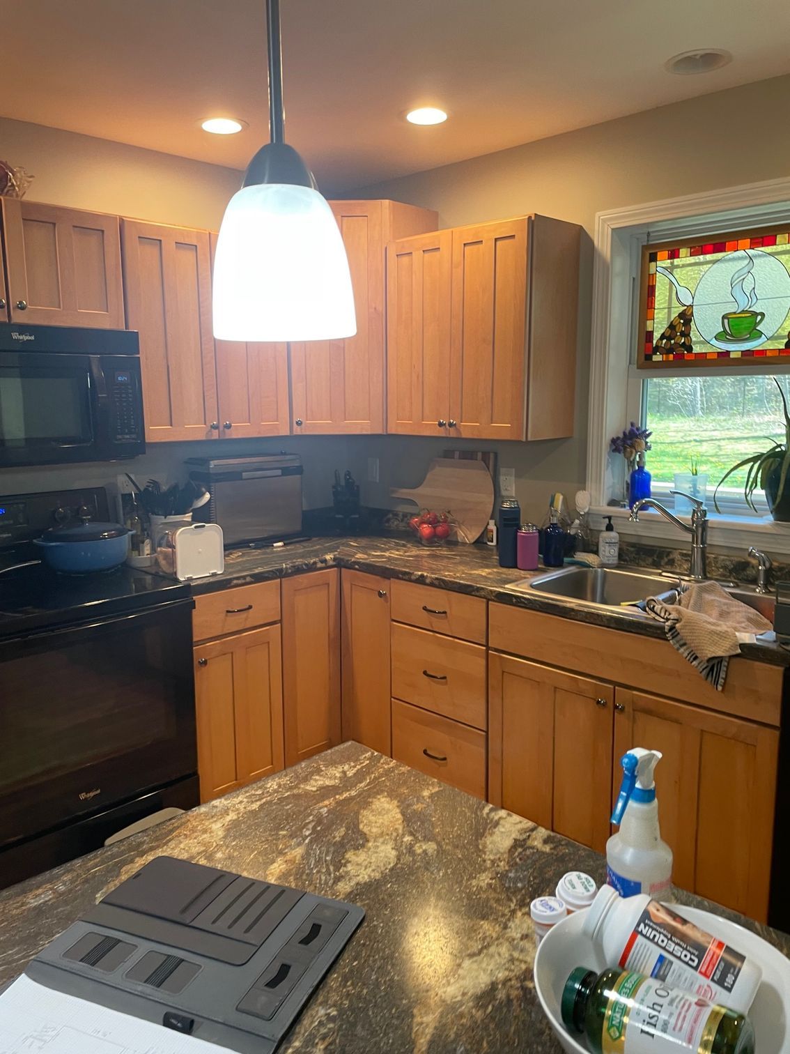 Cluttered kitchen with wooden cabinets, black appliances, hanging light, and sink by a window.