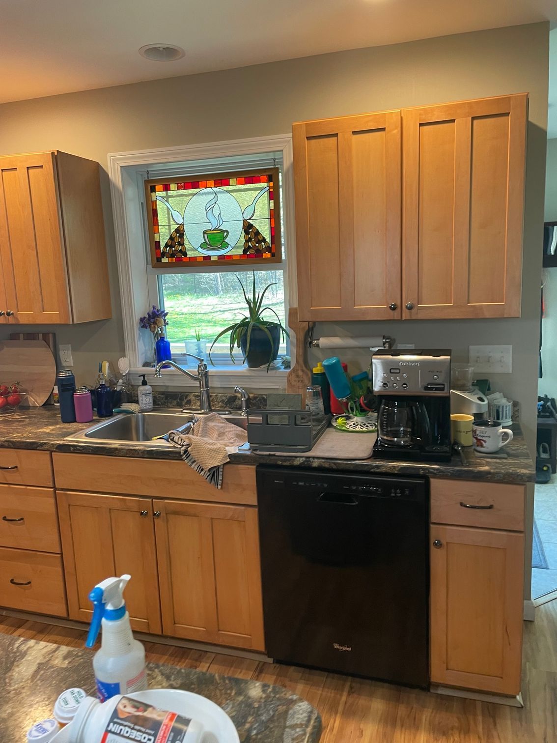 Kitchen with wooden cabinets, black dishwasher, sink under a stained-glass window, and cleaning supplies on the counter