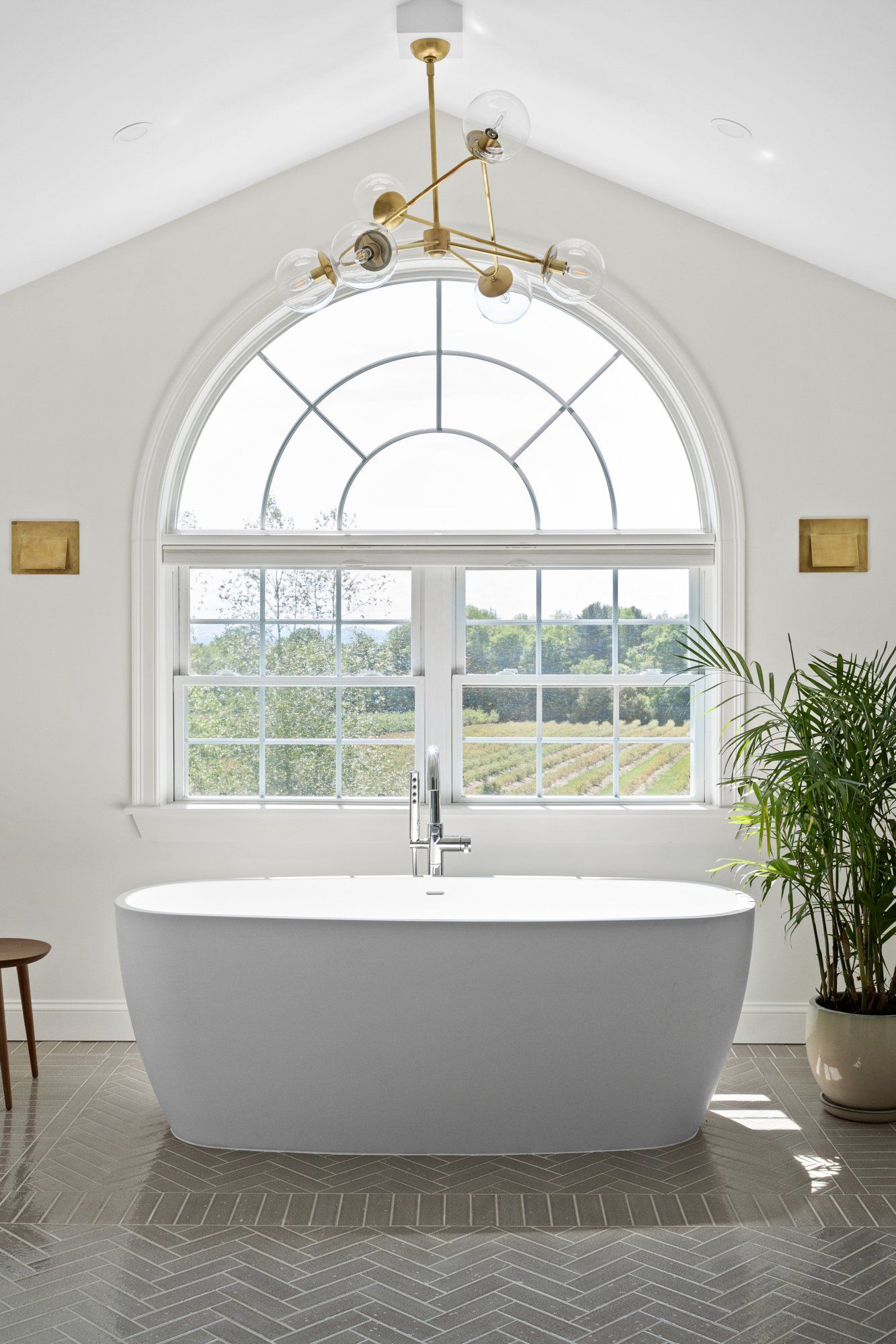 Bright bathroom with freestanding tub beneath a large arched window and gold chandelier