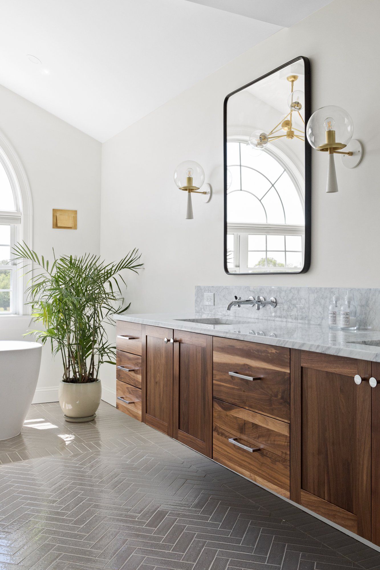 Bright bathroom with double vanity, gray tile, arched mirror, wall sconces, and a potted plant near a tub.