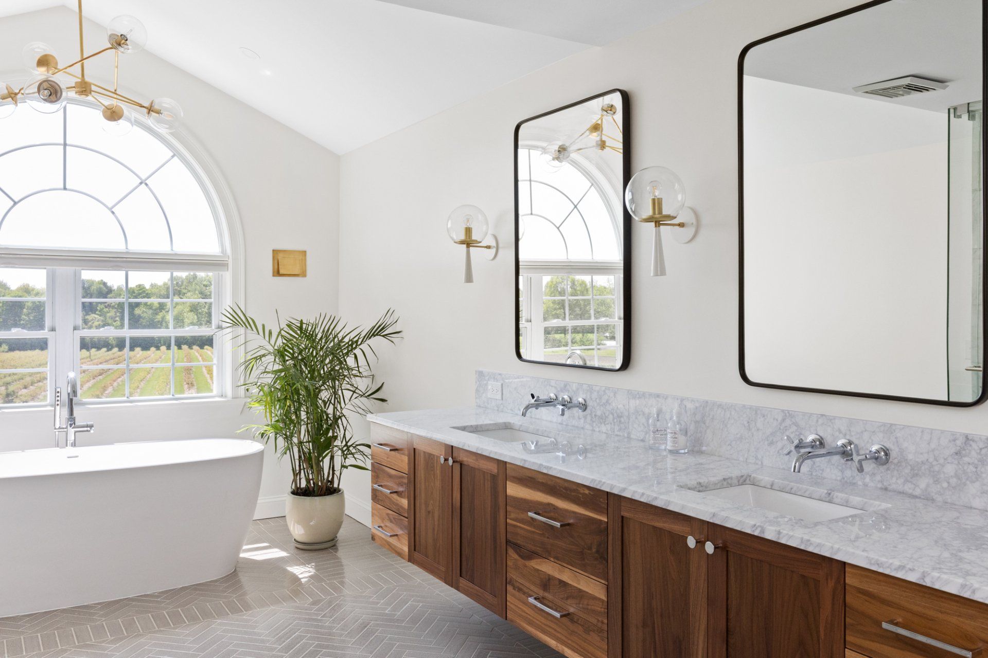 Bright bathroom with freestanding tub, double vanity, mirrors, and large arched window with potted plant