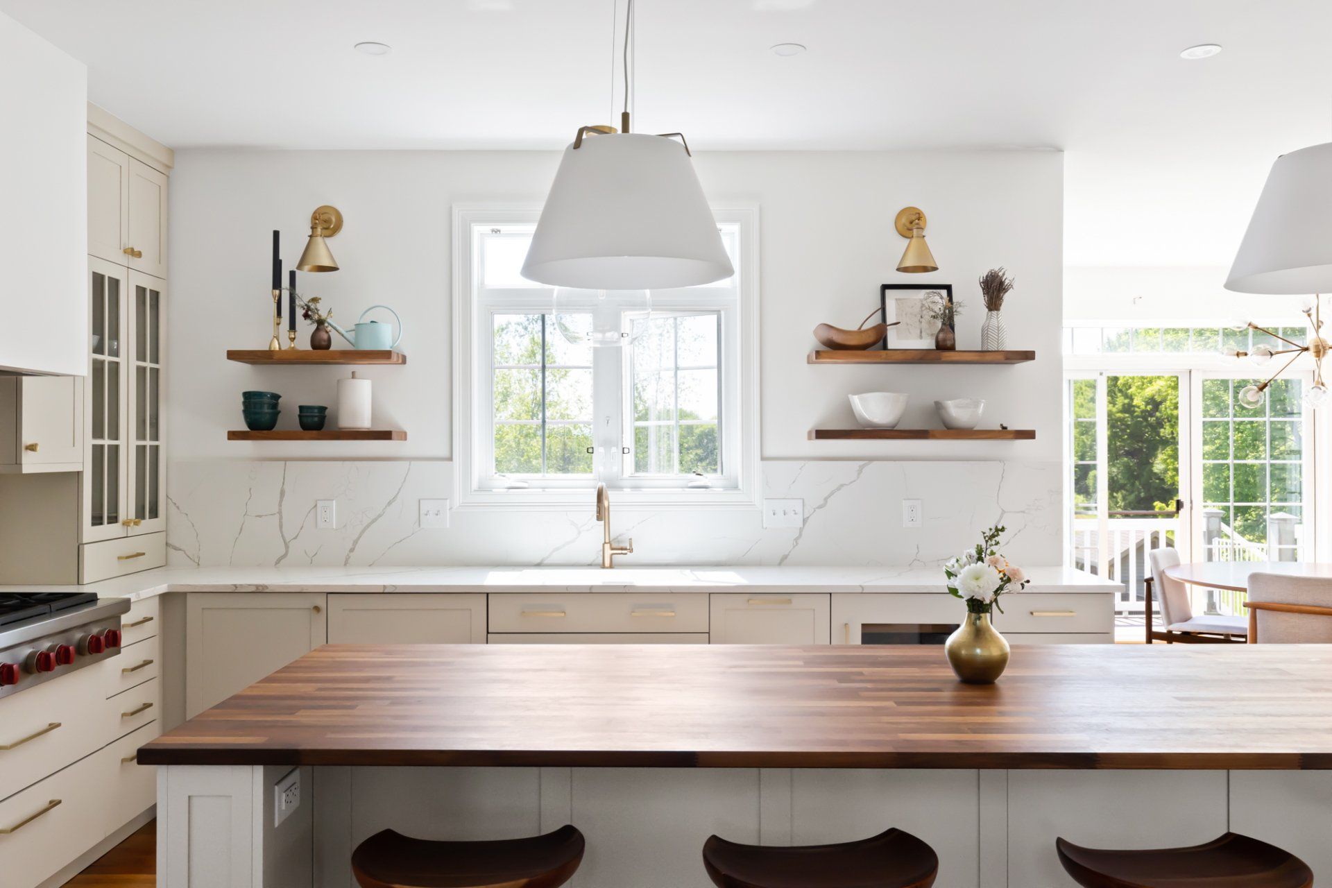 Bright white kitchen with marble island, wood countertops, pendant lights, and wall shelves by a window