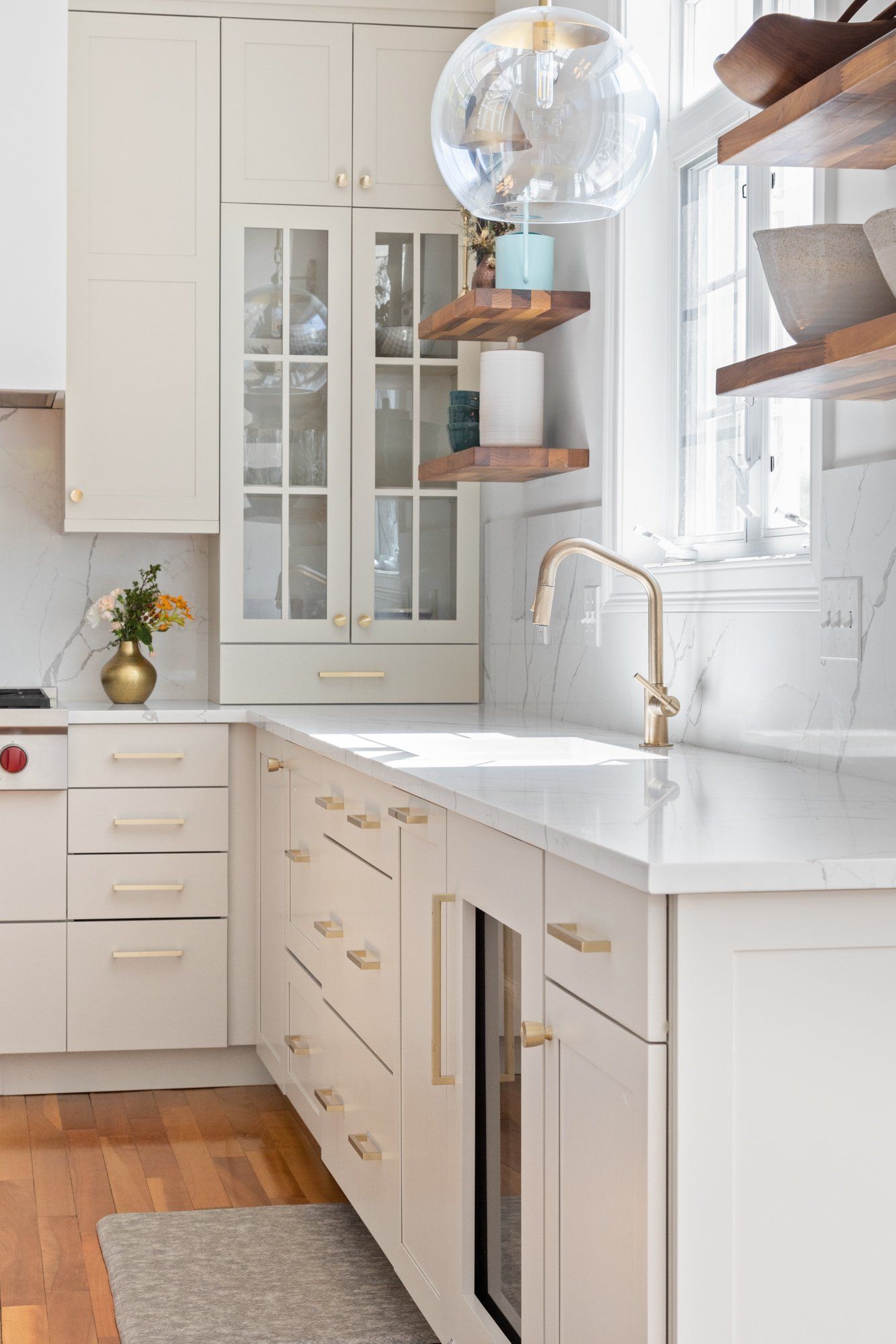 Bright white kitchen with gold fixtures, wood floors, glass cabinet doors, and a hanging globe light.