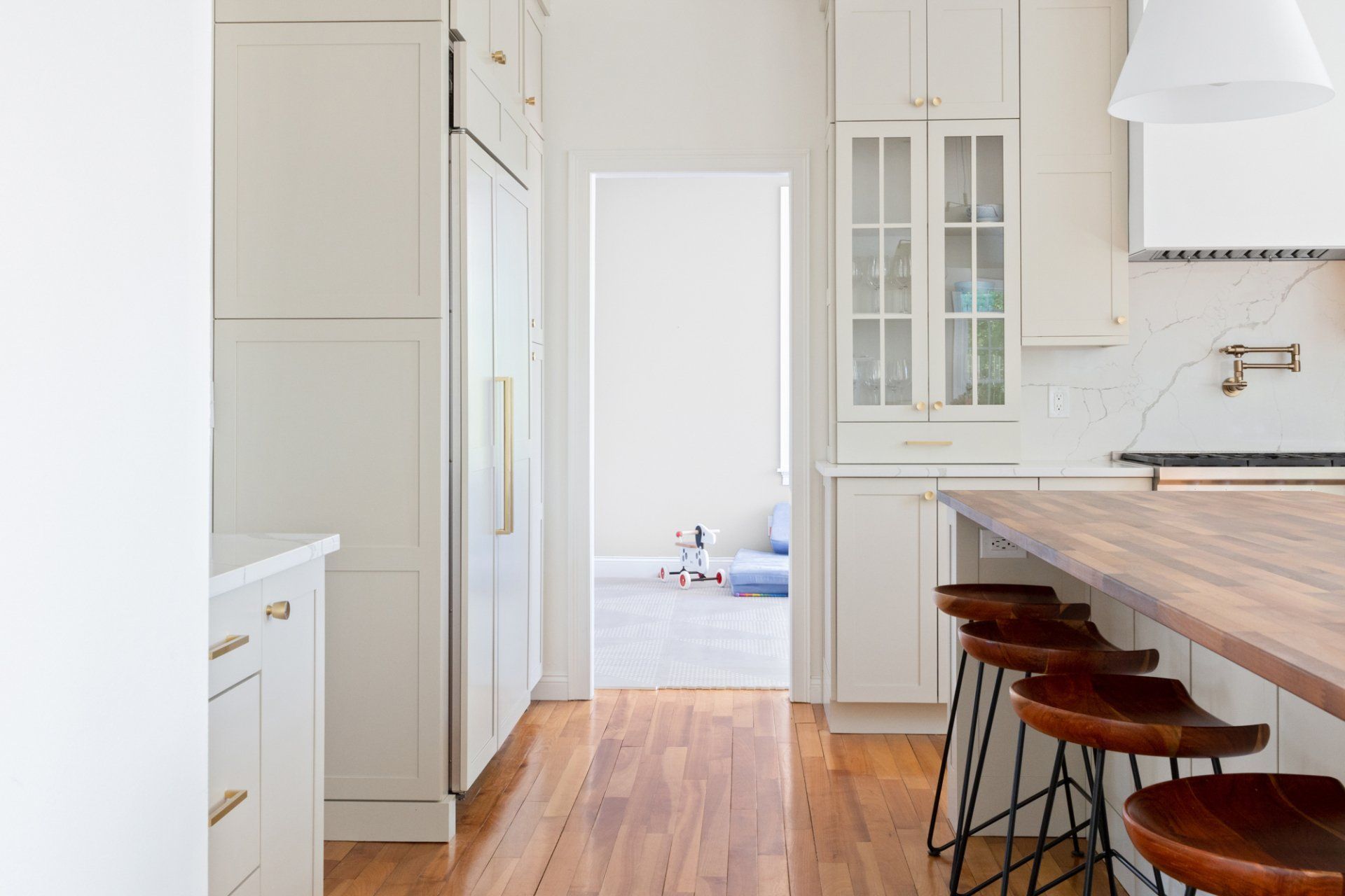 Bright kitchen with white cabinets, wood floor, and a marble island with stools, leading to a doorway.