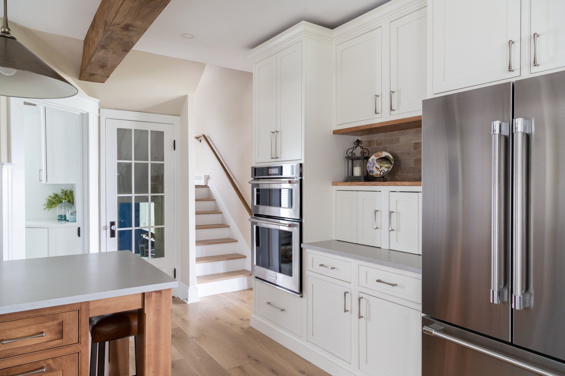 Bright white kitchen with stainless steel refrigerator, built-in oven, and staircase leading upstairs