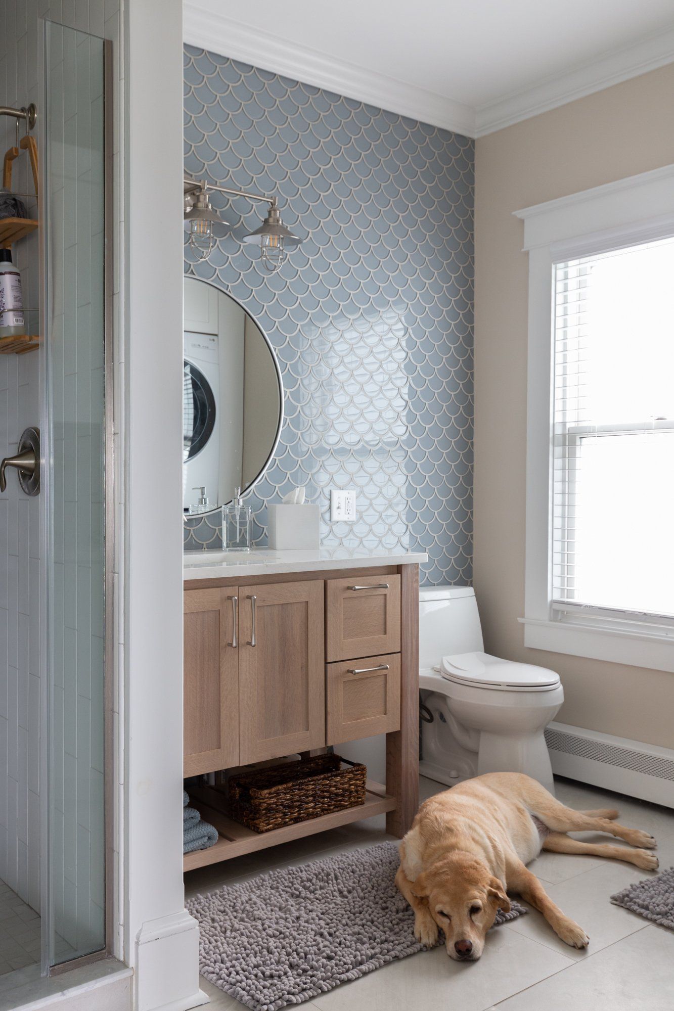 Bathroom with patterned tile, wooden vanity, round mirror, and a dog lying on a rug by the window.