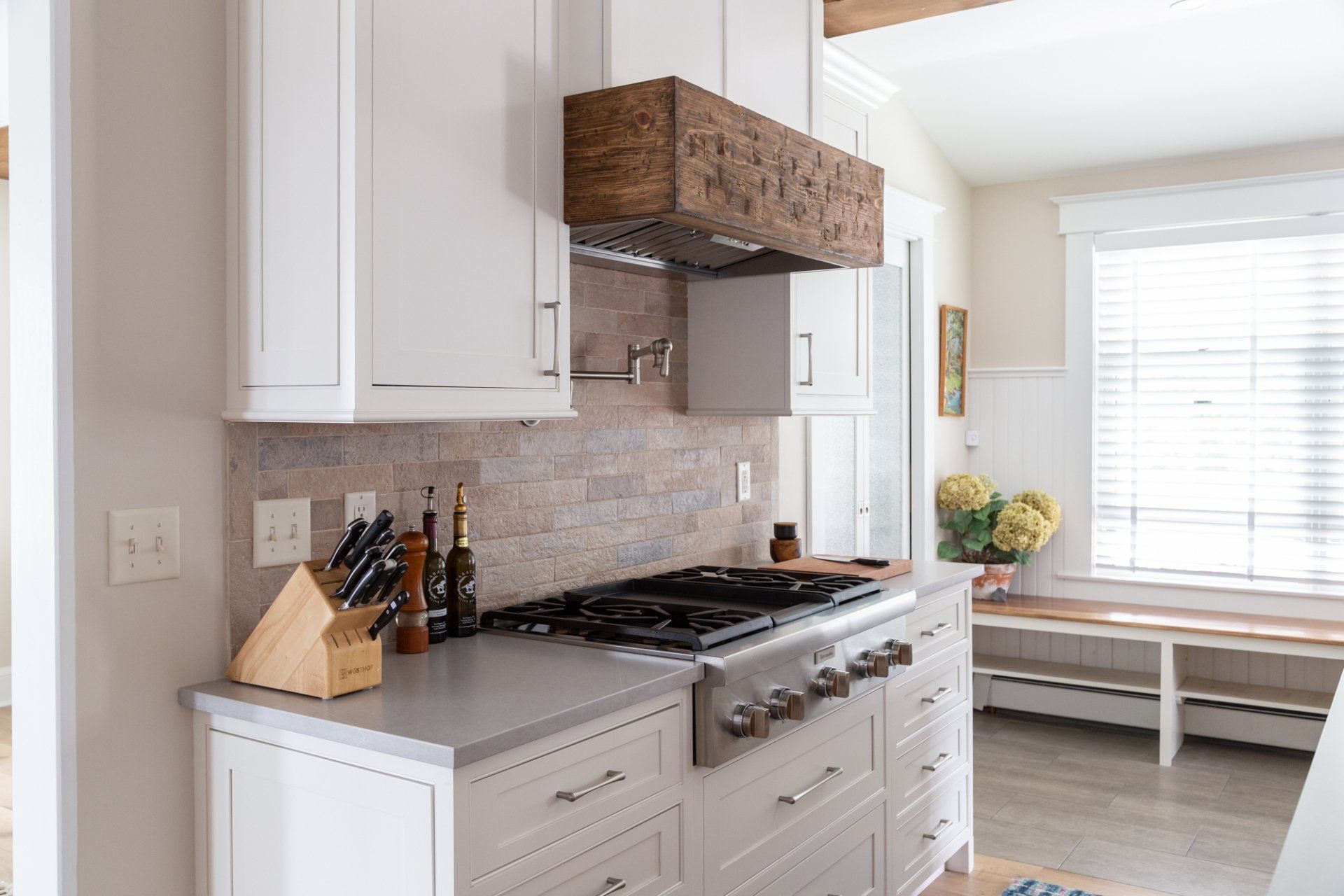 Bright white kitchen with stainless stove, wooden hood, and window seat by a sunlit window