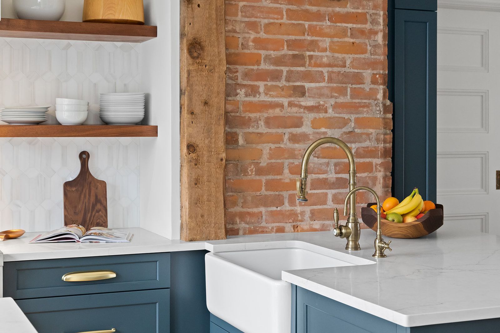 Bright kitchen with white farmhouse sink, brass faucet, blue cabinets, brick wall, and open shelves with dishes