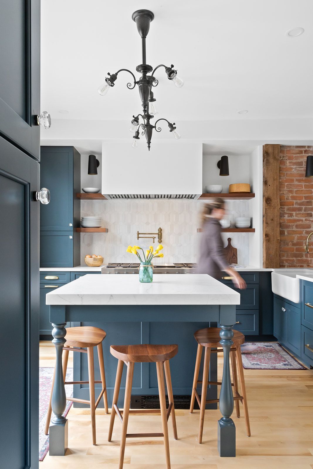 Modern kitchen with blue island, wooden stools, brick wall, and a person moving in the background