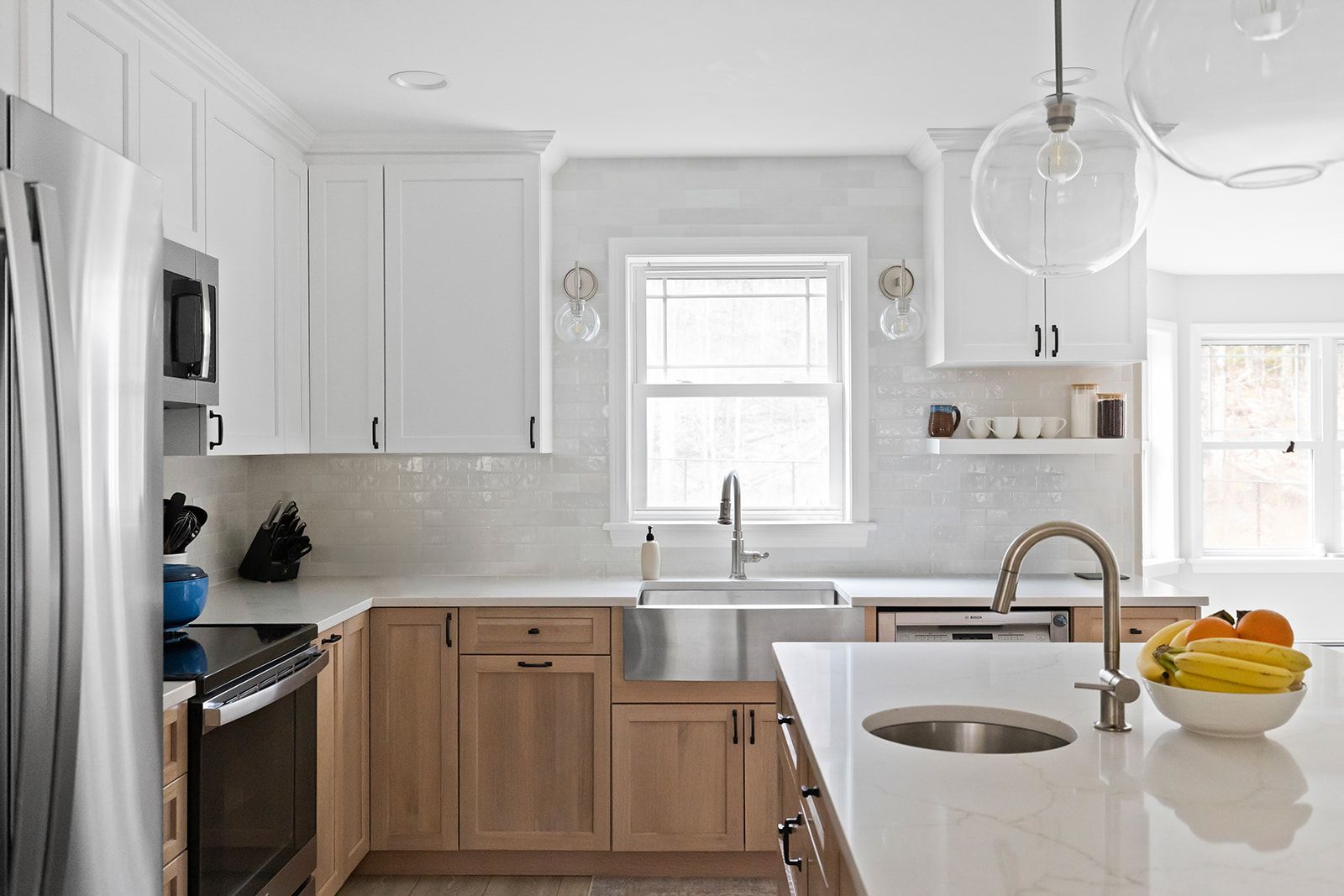 Bright modern white kitchen with wood cabinets, island sink, and bowl of fruit near a window.