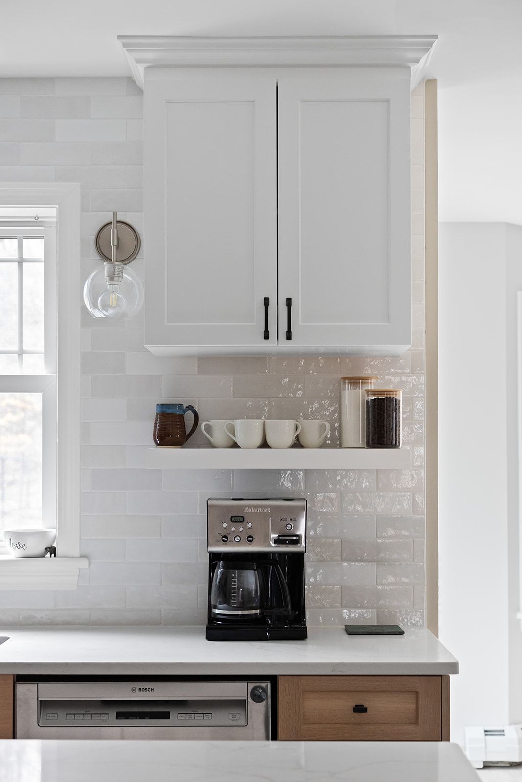 White kitchen cabinets above countertop with coffee maker, shelves, and stainless dishwasher beneath window.