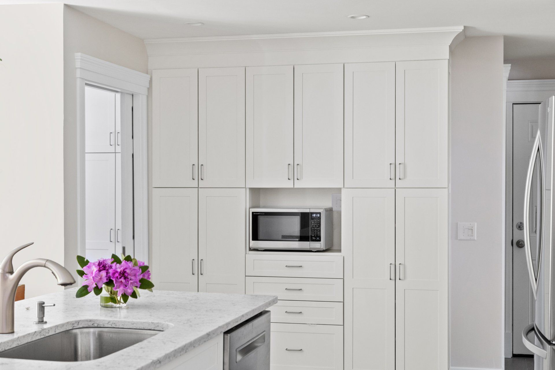 Bright white kitchen with island sink, microwave, cabinets, and stainless steel refrigerator.