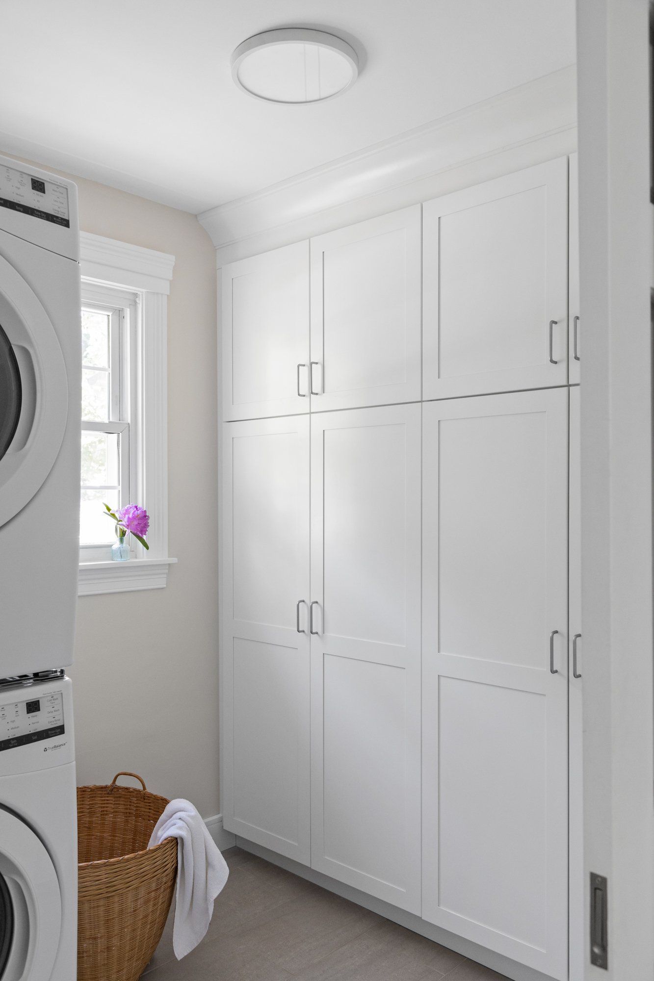 Bright laundry room with white cabinets, stacked washer and dryer, window, and wicker basket