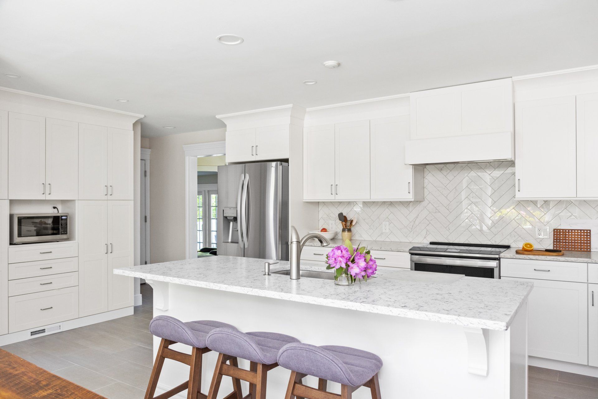 Bright white modern kitchen with island, marble counters, stainless fridge, and purple barstools
